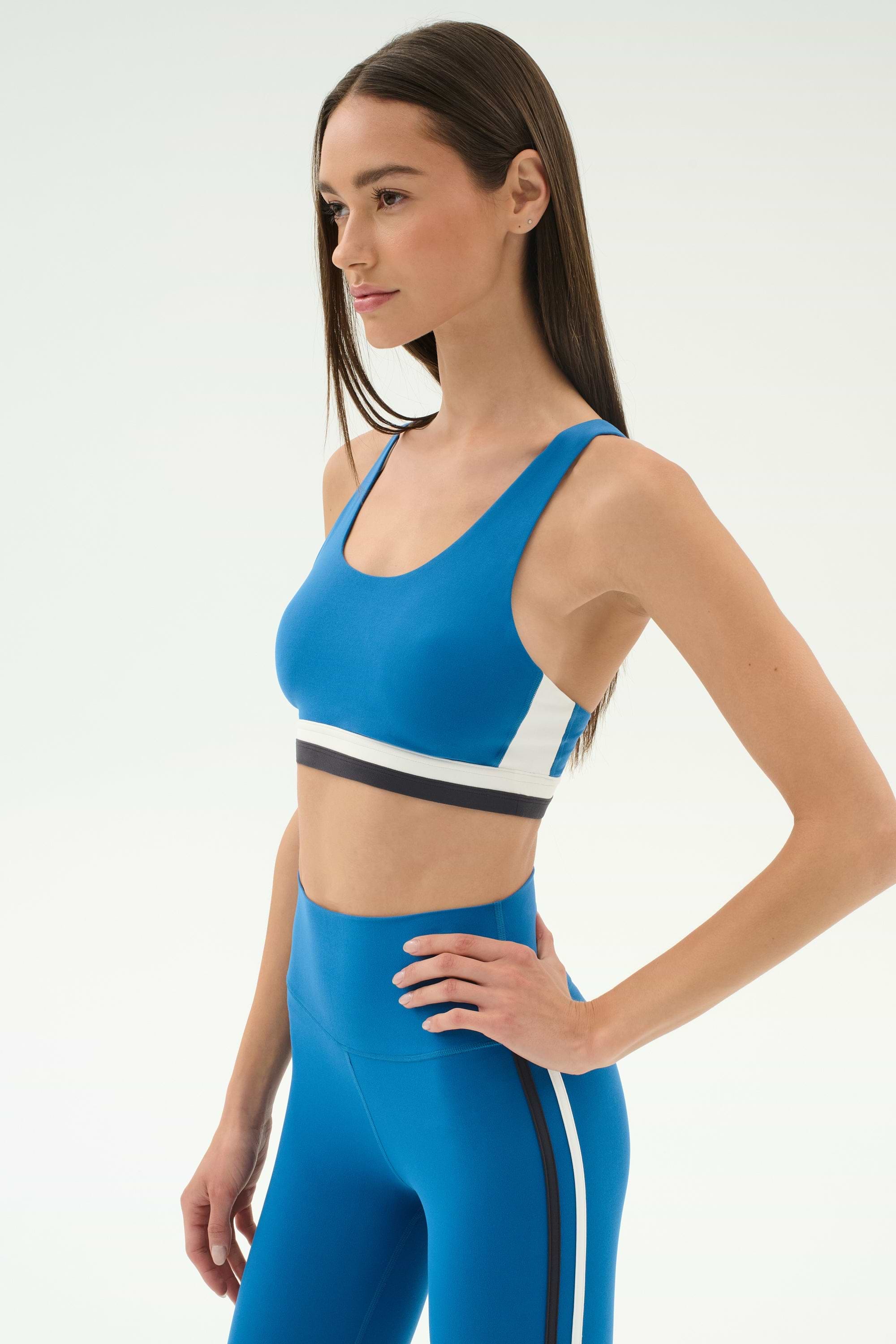 A woman wearing the SPLITS59 Maci Airweight Bra in Stone Blue/Graphite and matching high-waisted leggings with black and white stripes stands with one hand on her hip against a plain background.