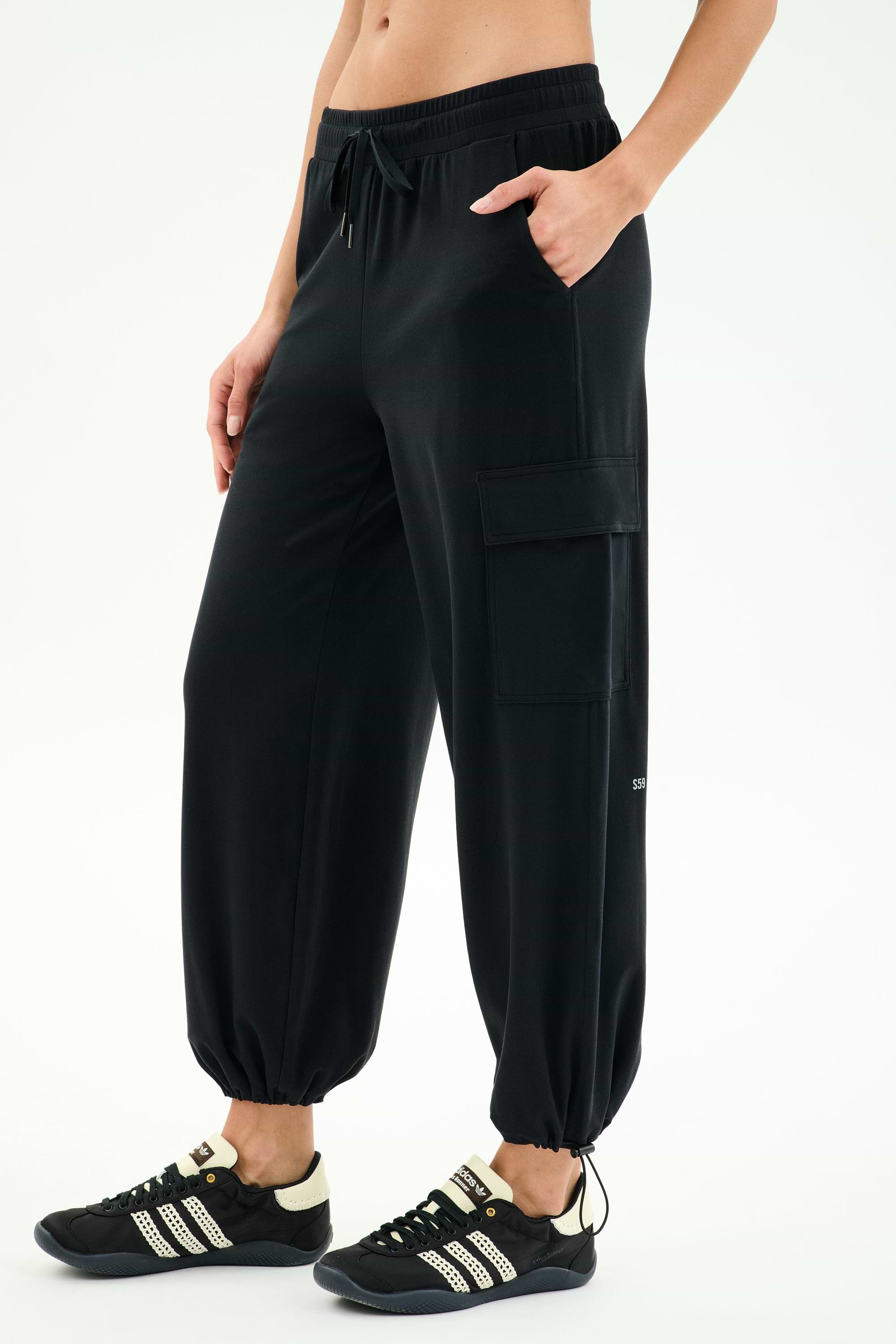 A person wears the SPLITS59 Nico Airweight Cargo Pant in black, paired with black sneakers with white stripes and a cropped black top, standing with one hand in a pocket for effortless comfort.