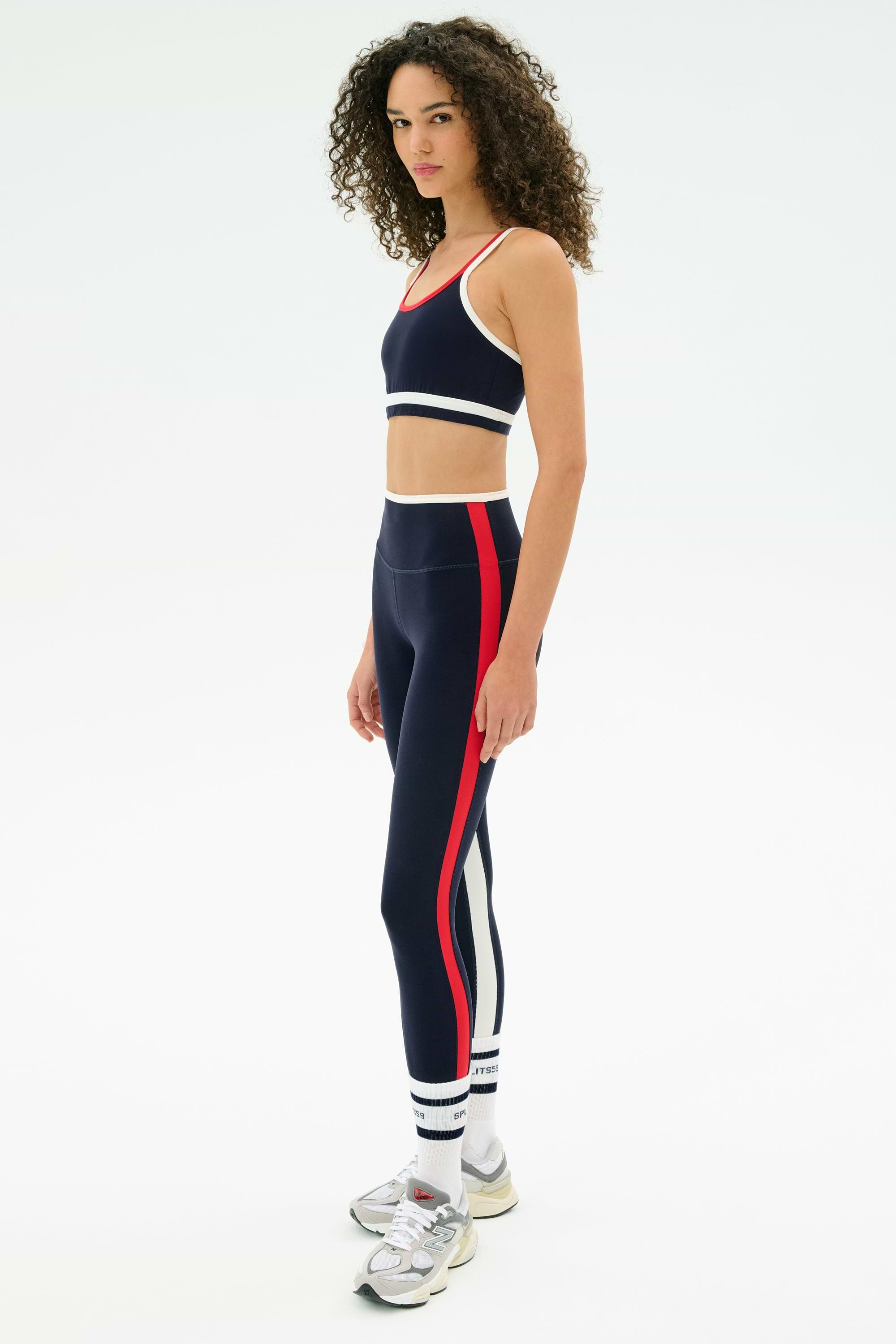 A woman models the SPLITS59 Eva Rigor Double Strap Bra in Indigo/White with white sneakers and curly hair, standing against a plain white background—showcasing sporty comfort and confidence for workouts or daily wear.