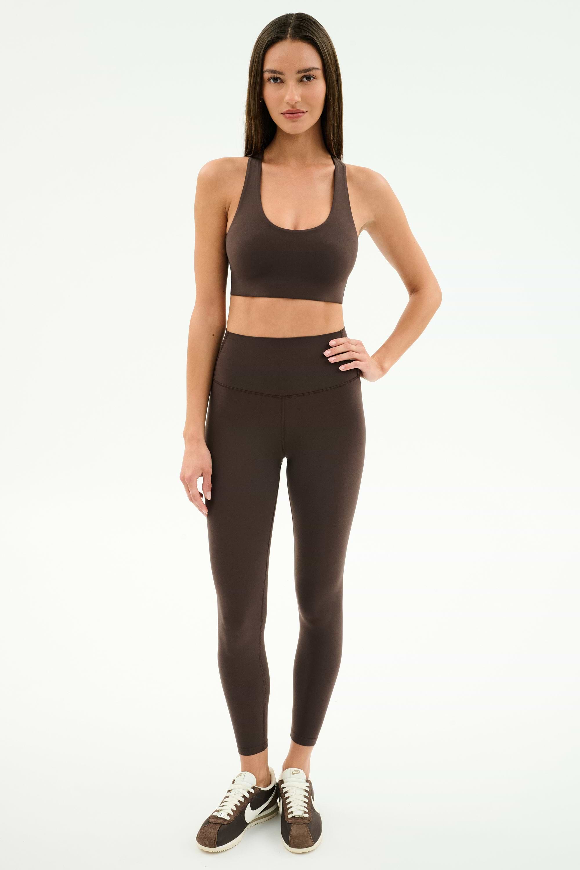 A woman stands facing forward in SPLITS59’s Airweight High Waist Legging in Dark Chocolate with a matching sports bra and black-and-white sneakers, set against a plain white background.