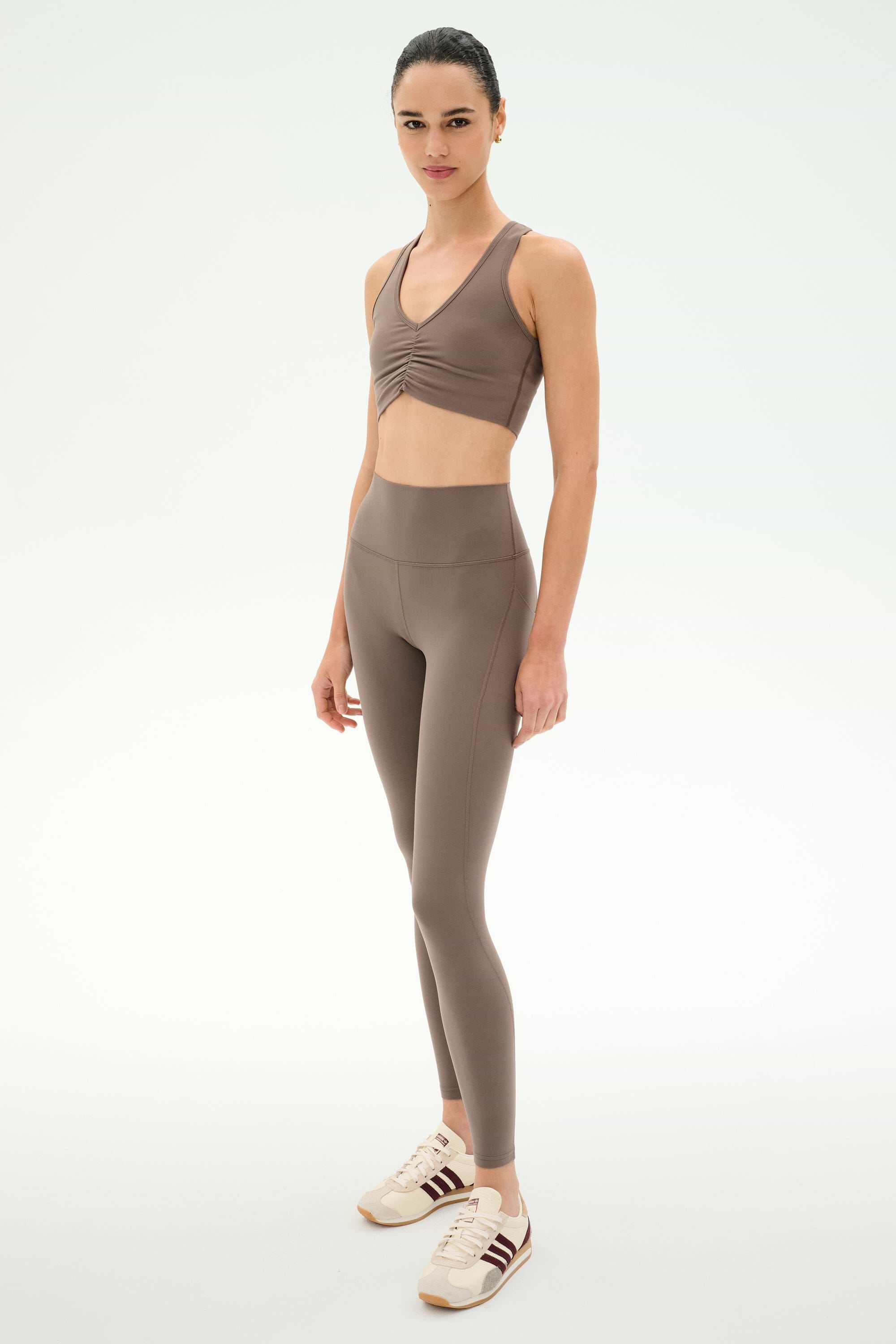 A woman stands in the SPLITS59 Airweight Bra With Ruching in Lentil and high-waisted leggings, both crafted from buttery-soft stretch fabric, paired with white sneakers against a plain white background.