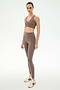 A woman stands in SPLITS59’s Airweight High Waist 7/8 Pocket Legging in Lentil, paired with a taupe spandex sports bra and cream and brown sneakers, against a plain white background.