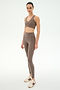 A woman stands in SPLITS59’s Airweight High Waist 7/8 Pocket Legging in Lentil, paired with a taupe spandex sports bra and cream and brown sneakers, against a plain white background.