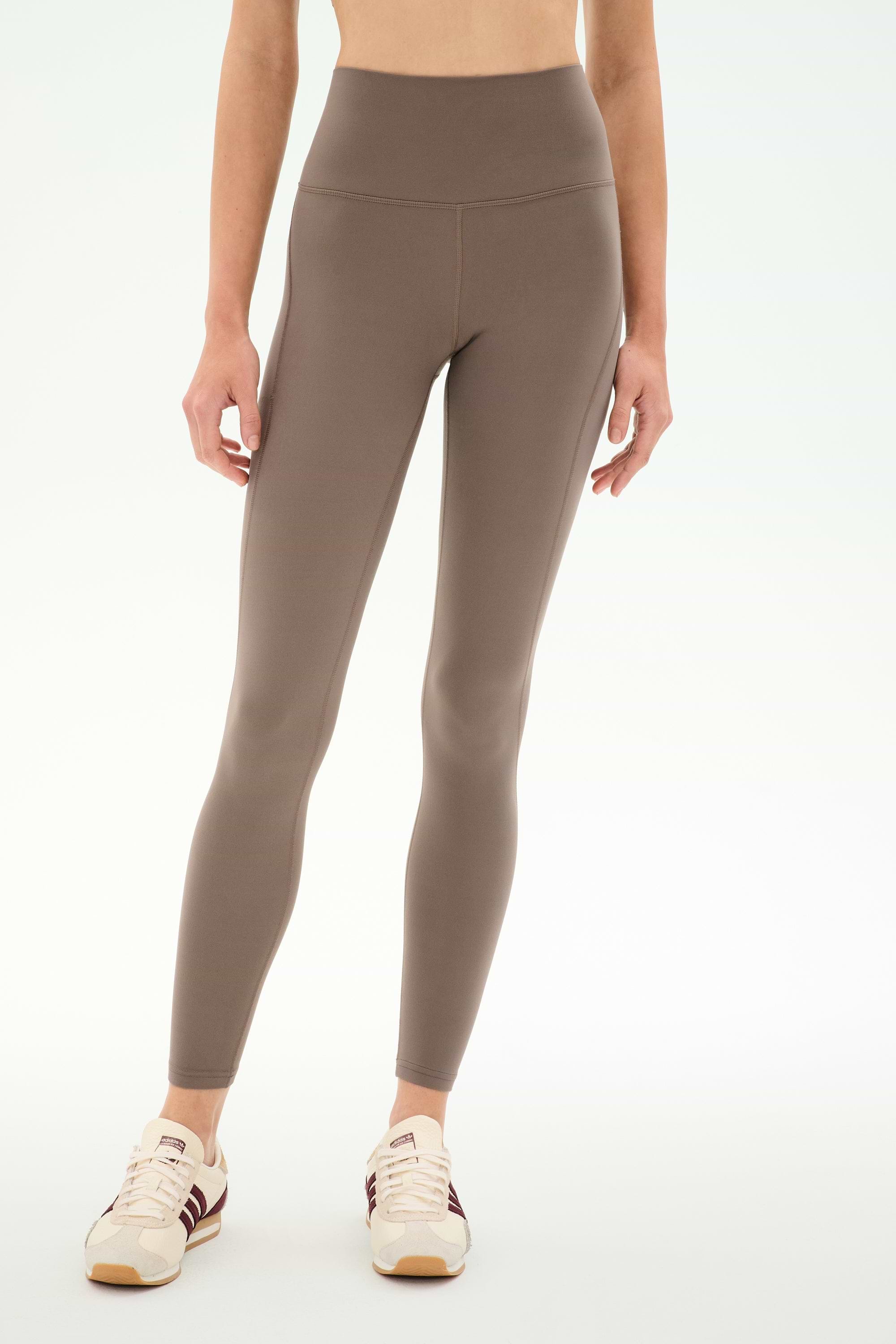 A person wearing SPLITS59′s Airweight High Waist 7/8 Pocket Legging in Lentil, paired with cream sneakers featuring brown accents, stands against a plain white background.
