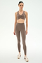 A person stands in a neutral pose wearing the SPLITS59 Airweight Bra With Ruching in Lentil, paired with matching high-waisted leggings made from stretch fabric and white sneakers, set against a plain light background to showcase modern athletic wear.