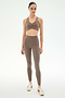 A woman stands facing forward against a plain white background wearing SPLITS59′s Airweight High Waist 7/8 Pocket Legging in Lentil, paired with a matching taupe nylon-spandex sports bra and white sneakers.