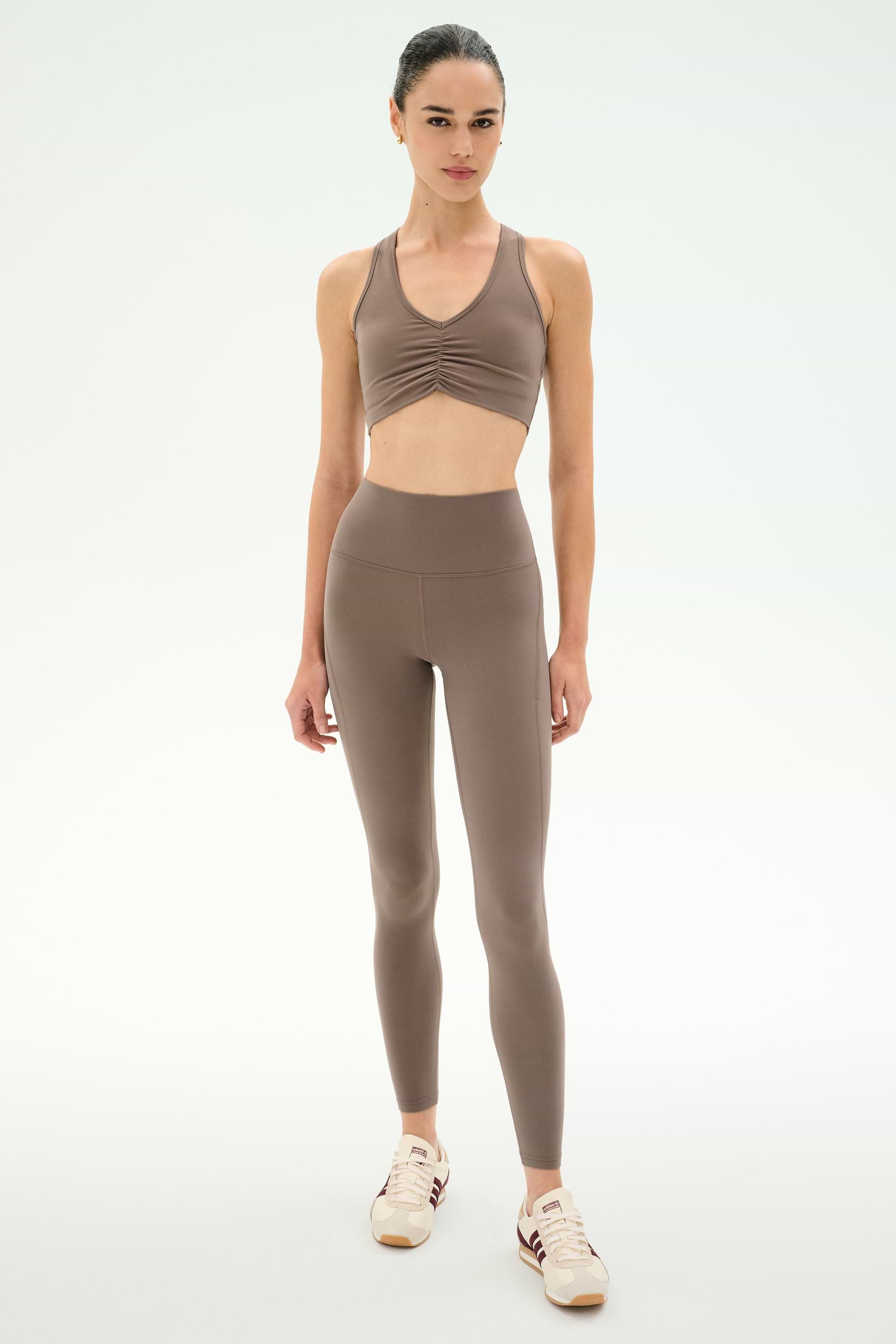 A woman stands facing forward against a plain white background wearing SPLITS59′s Airweight High Waist 7/8 Pocket Legging in Lentil, paired with a matching taupe nylon-spandex sports bra and white sneakers.