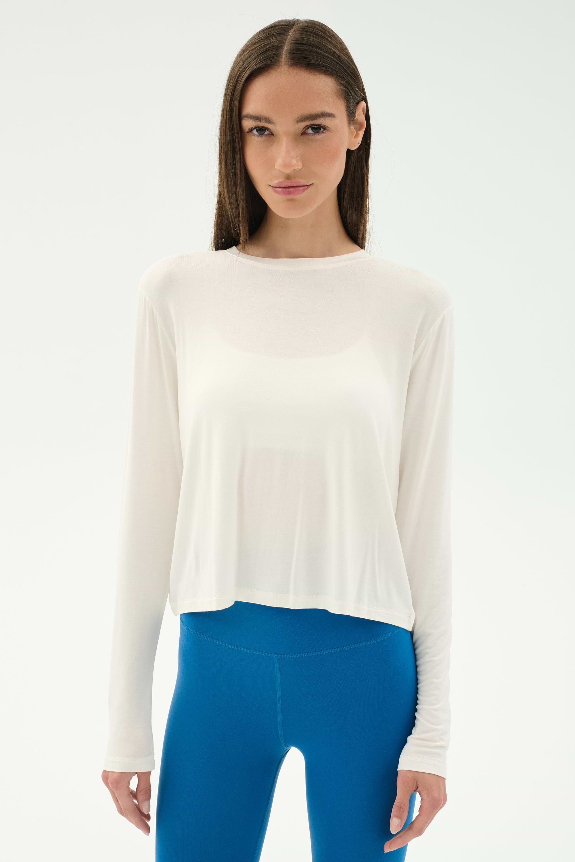A woman models the SPLITS59 Djuna Long Sleeve - White, paired with bright blue leggings, standing against a plain light background, ready for gym workouts.
