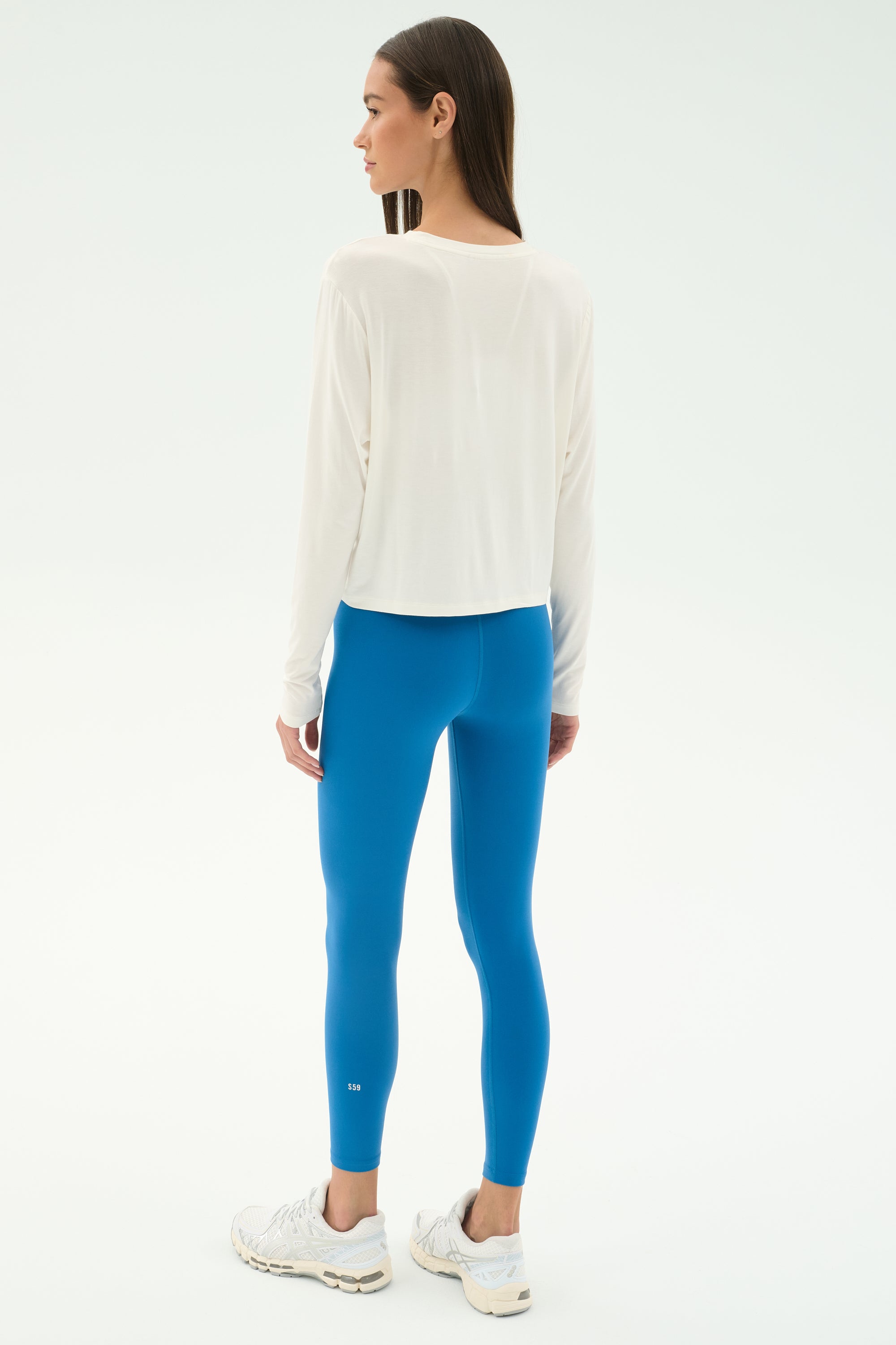 A woman faces away, wearing the SPLITS59 Djuna Long Sleeve - White, bright blue leggings, and white sneakers against a plain background—ideal for gym or outdoor workouts.