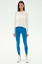A woman stands facing forward against a plain white background, wearing the SPLITS59 Djuna Long Sleeve - White, bright blue leggings, and white athletic shoes—ideal attire for gym workouts.