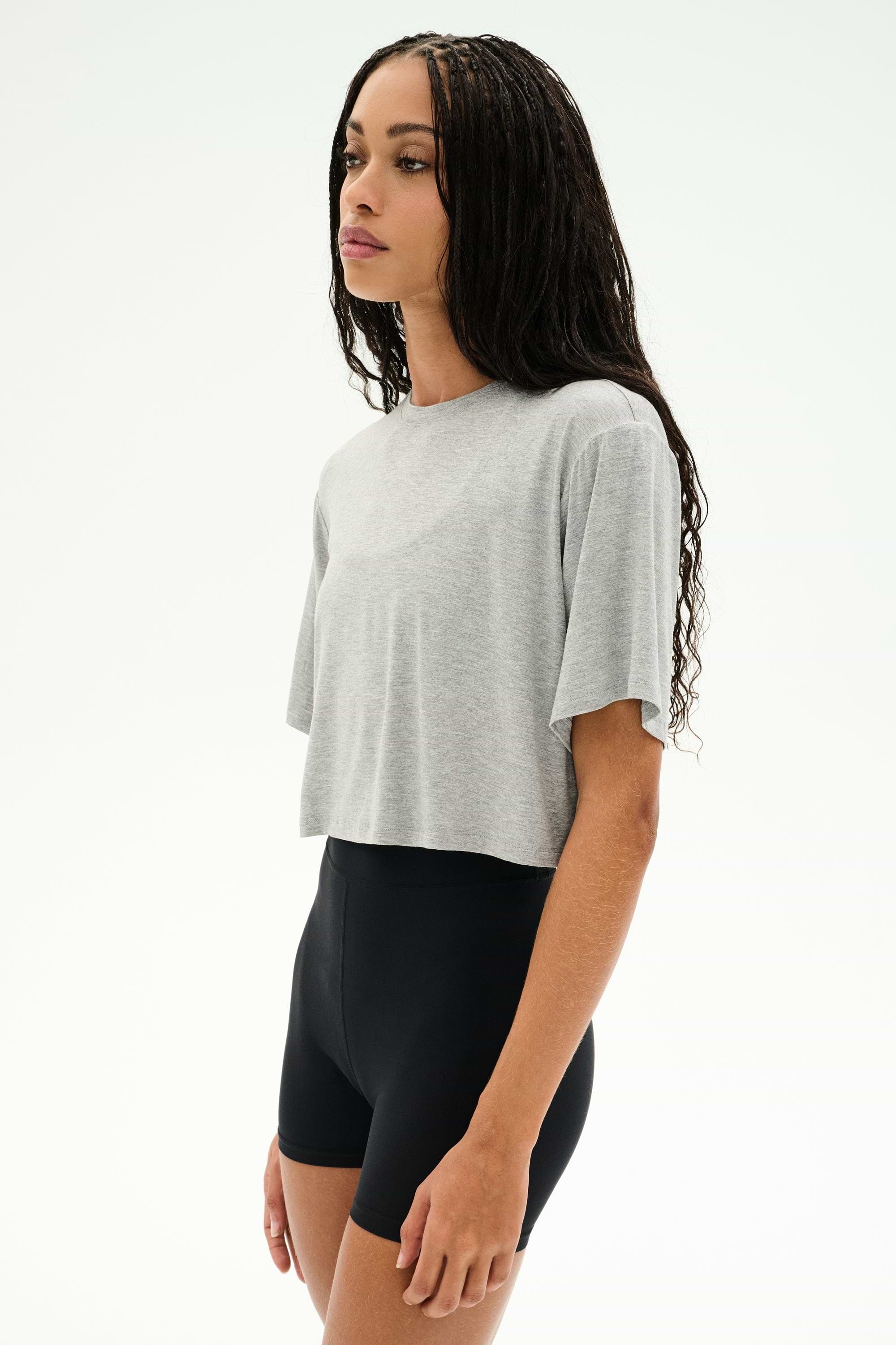 A woman wears the SPLITS59 Djuna Oversized Crop Jersey Tee in Heather Grey, paired with black high-waisted shorts, standing against a plain white background.