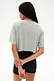 A person with long braided hair stands facing away, wearing the SPLITS59 Djuna Oversized Crop Jersey Tee in Heather Grey and black shorts, hands clasped behind their back against a plain light background.