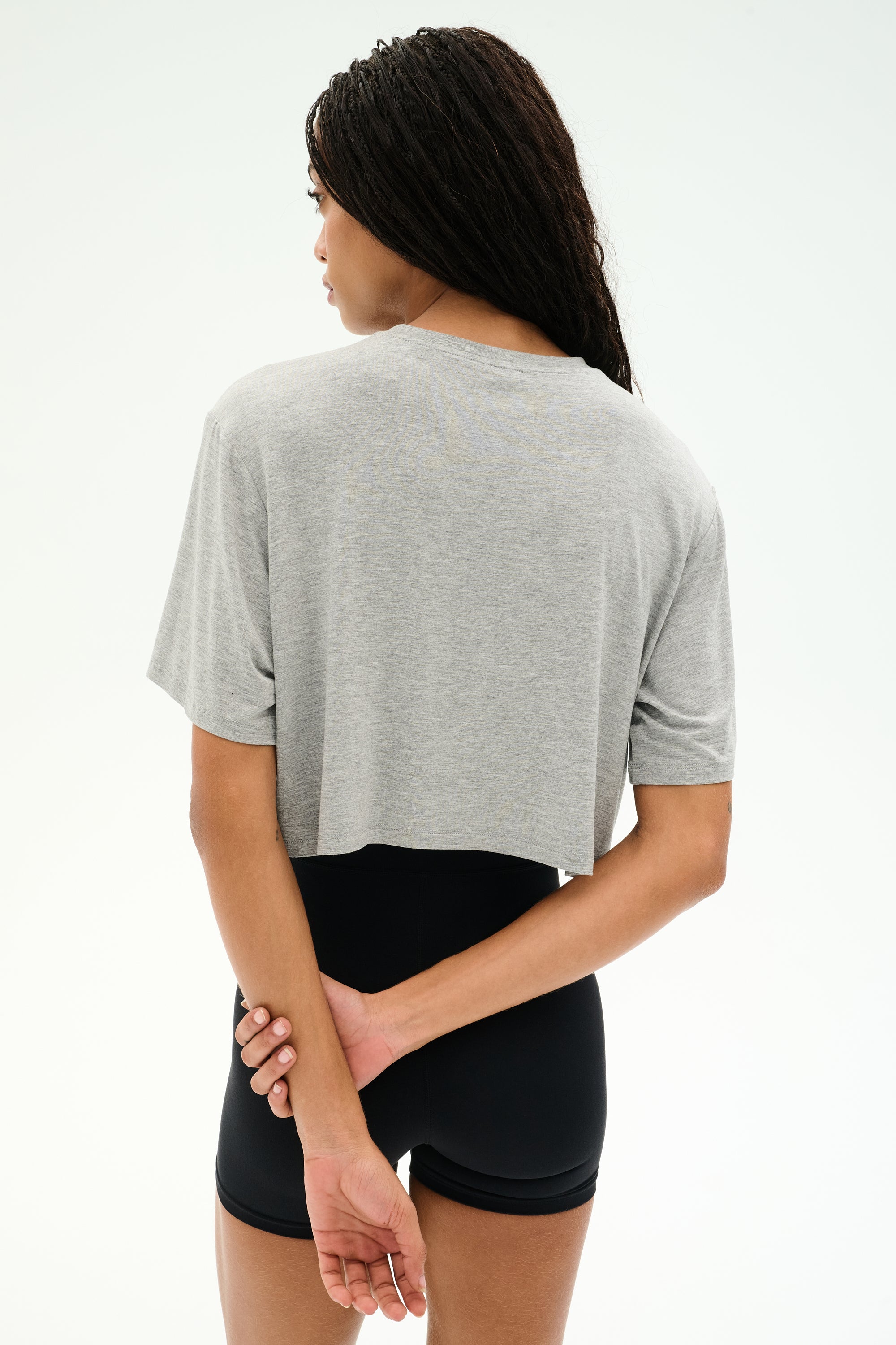 A person with long braided hair stands facing away, wearing the SPLITS59 Djuna Oversized Crop Jersey Tee in Heather Grey and black shorts, hands clasped behind their back against a plain light background.
