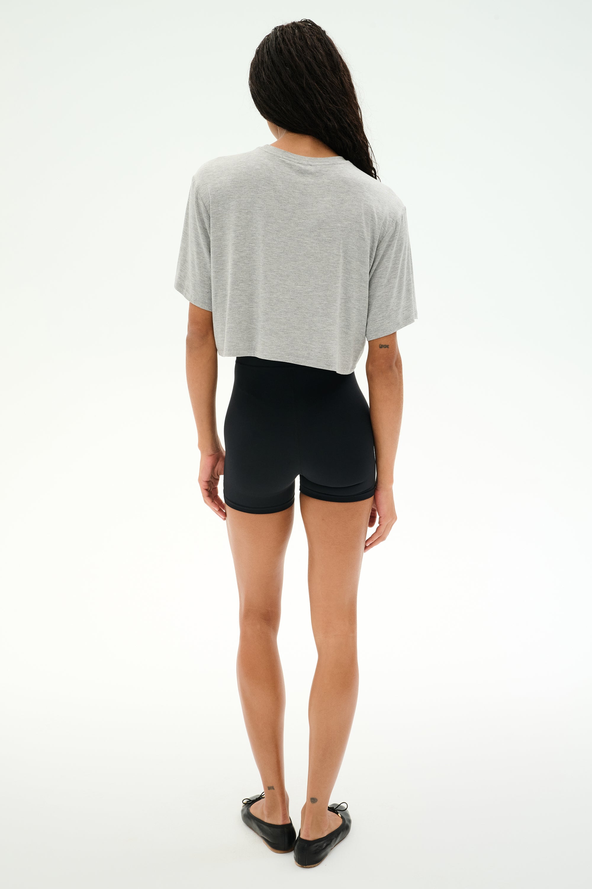 A person stands facing away against a white background, wearing SPLITS59's Djuna Oversized Crop Jersey Tee in Heather Grey, paired with black shorts and black flats.