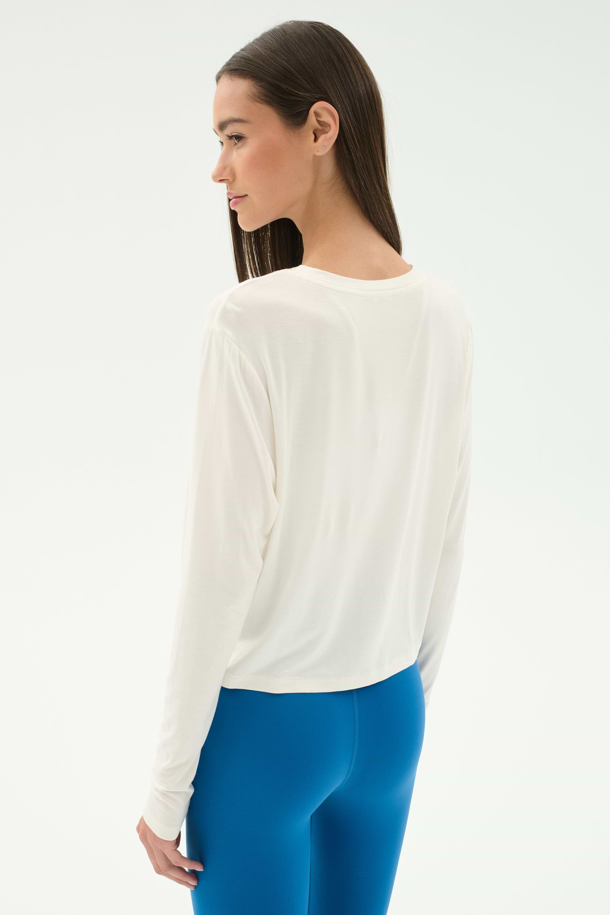 A woman stands sideways against a light background, wearing the SPLITS59 Djuna Long Sleeve in white and bright blue leggings.