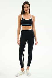 A woman faces forward in the SPLITS59 Eva Rigor Double Strap Bra in Black/Skylight, paired with high-waisted black leggings and white sneakers, against a plain white background.