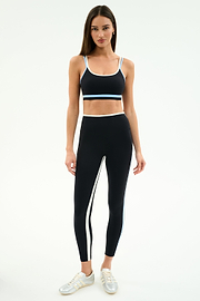A woman faces forward in the SPLITS59 Eva Rigor Double Strap Bra in Black/Skylight, paired with high-waisted black leggings and white sneakers, against a plain white background.