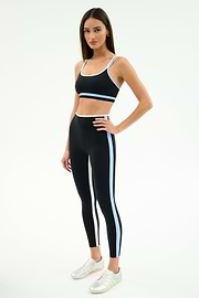 A woman wears the SPLITS59 Eva Rigor Double Strap Bra in Black/Skylight with matching leggings, white sneakers, and stands against a plain white background.