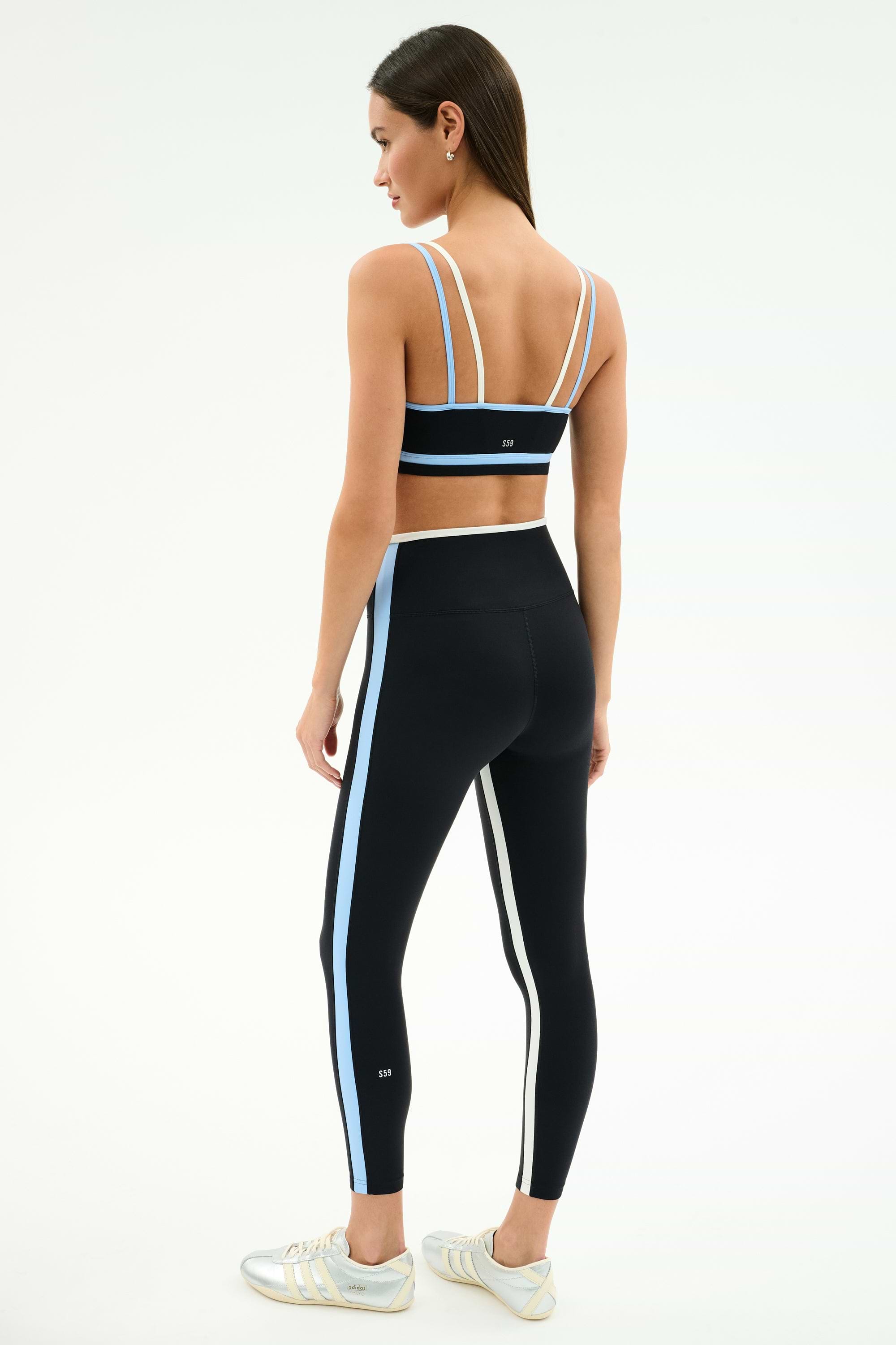 A woman stands sideways on a white background, wearing SPLITS59's Sam High Waist Rigor 7/8 Leggings in Black/Skylight with white and blue side stripes, paired with a matching black run tight sports bra.