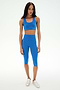 A woman stands facing forward against a plain white background, wearing the SPLITS59 Airweight High Waist 16" Capri Legging in Classic Blue, paired with a matching blue sports bra and white sneakers.