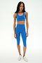 A woman stands facing forward against a plain white background, wearing the SPLITS59 Airweight High Waist 16" Capri Legging in Classic Blue, paired with a matching blue sports bra and white sneakers.