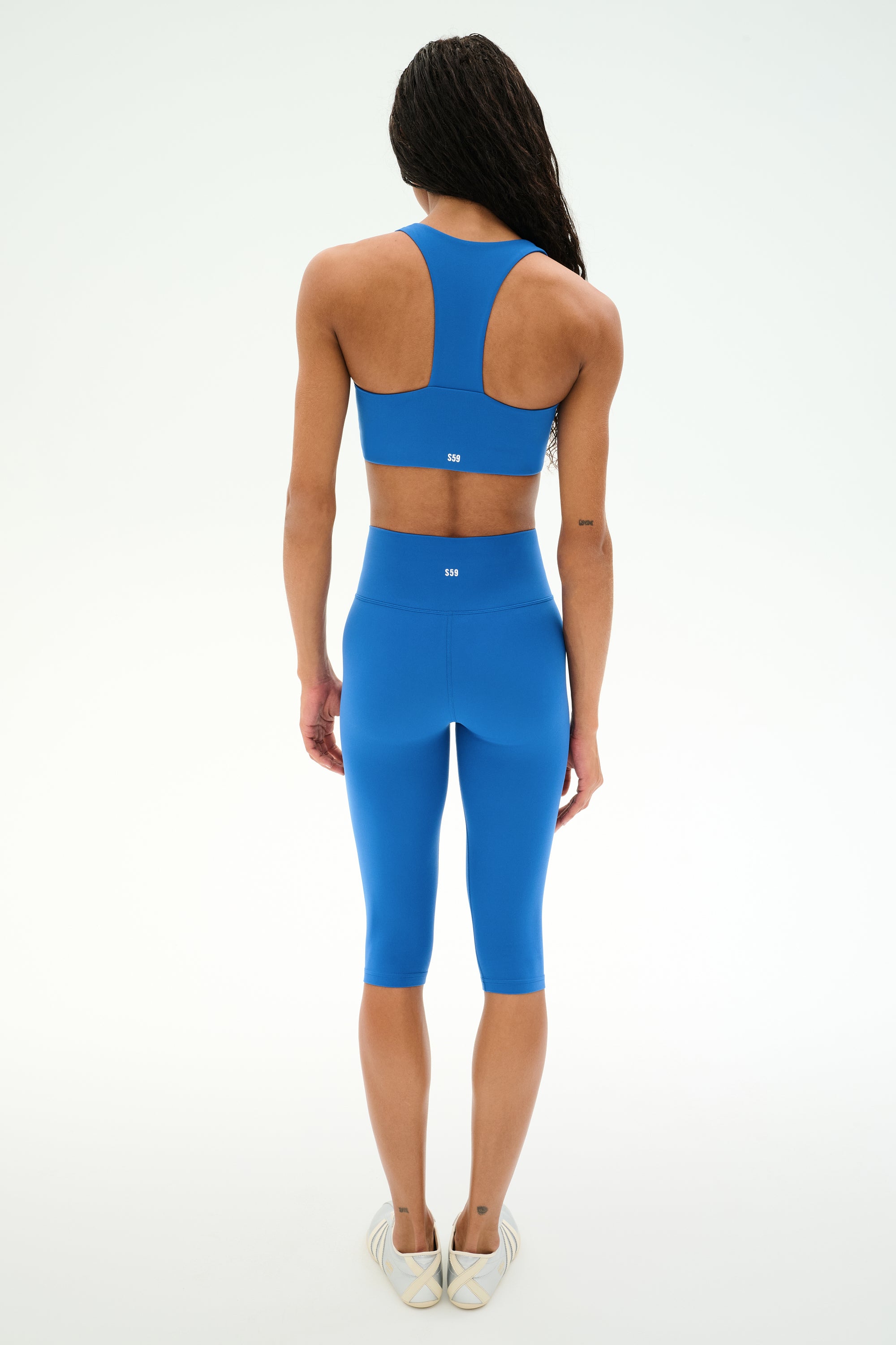 A person stands with their back to the camera, wearing the SPLITS59 Airweight High Waist 16" Capri Legging in Classic Blue, paired with a matching blue sports bra, against a plain white background.