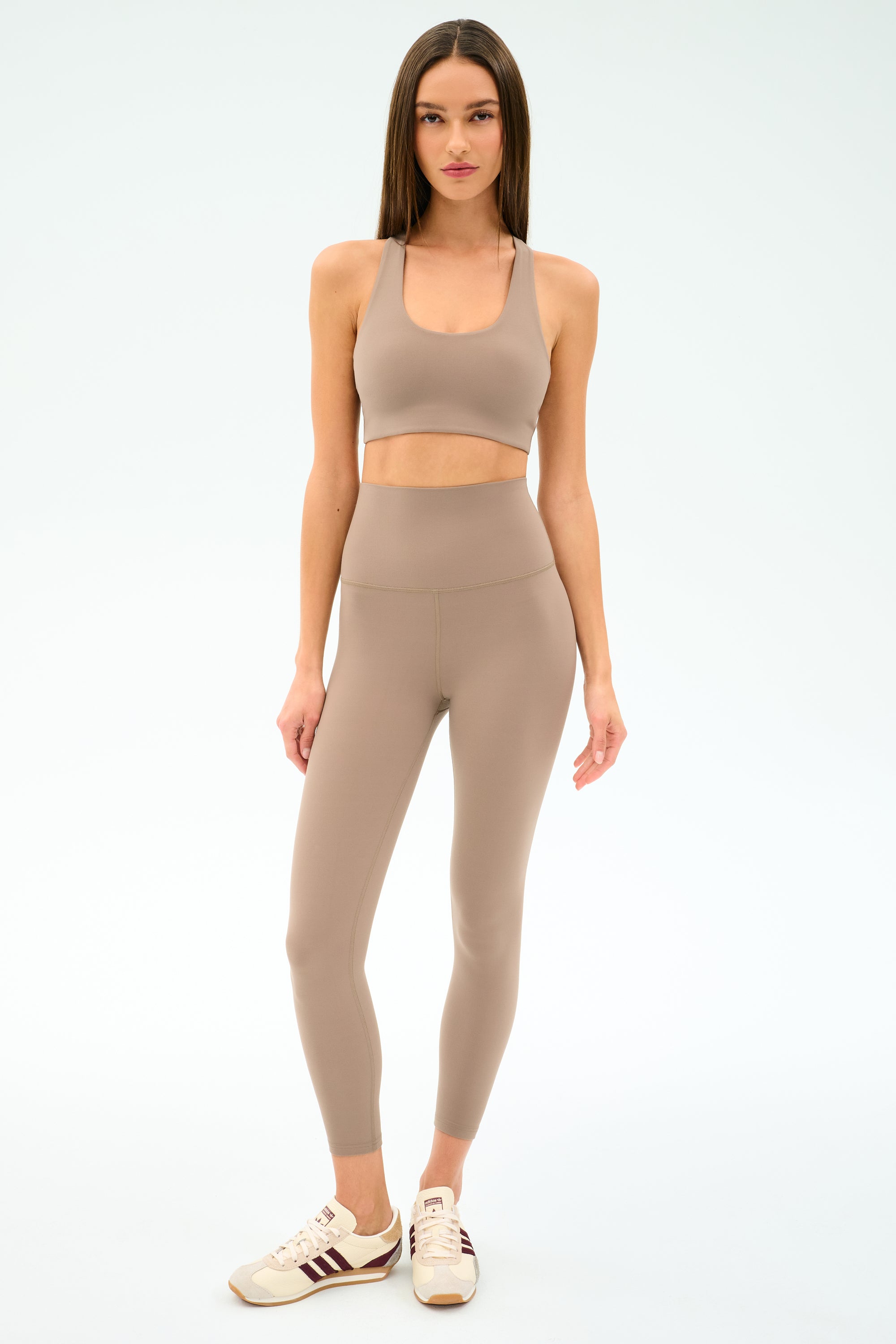 A woman stands against a plain background wearing the SPLITS59 Sara Airweight Bra in Latte, matching high-waisted leggings ideal for Pilates or hot yoga, and beige sneakers with maroon stripes.
