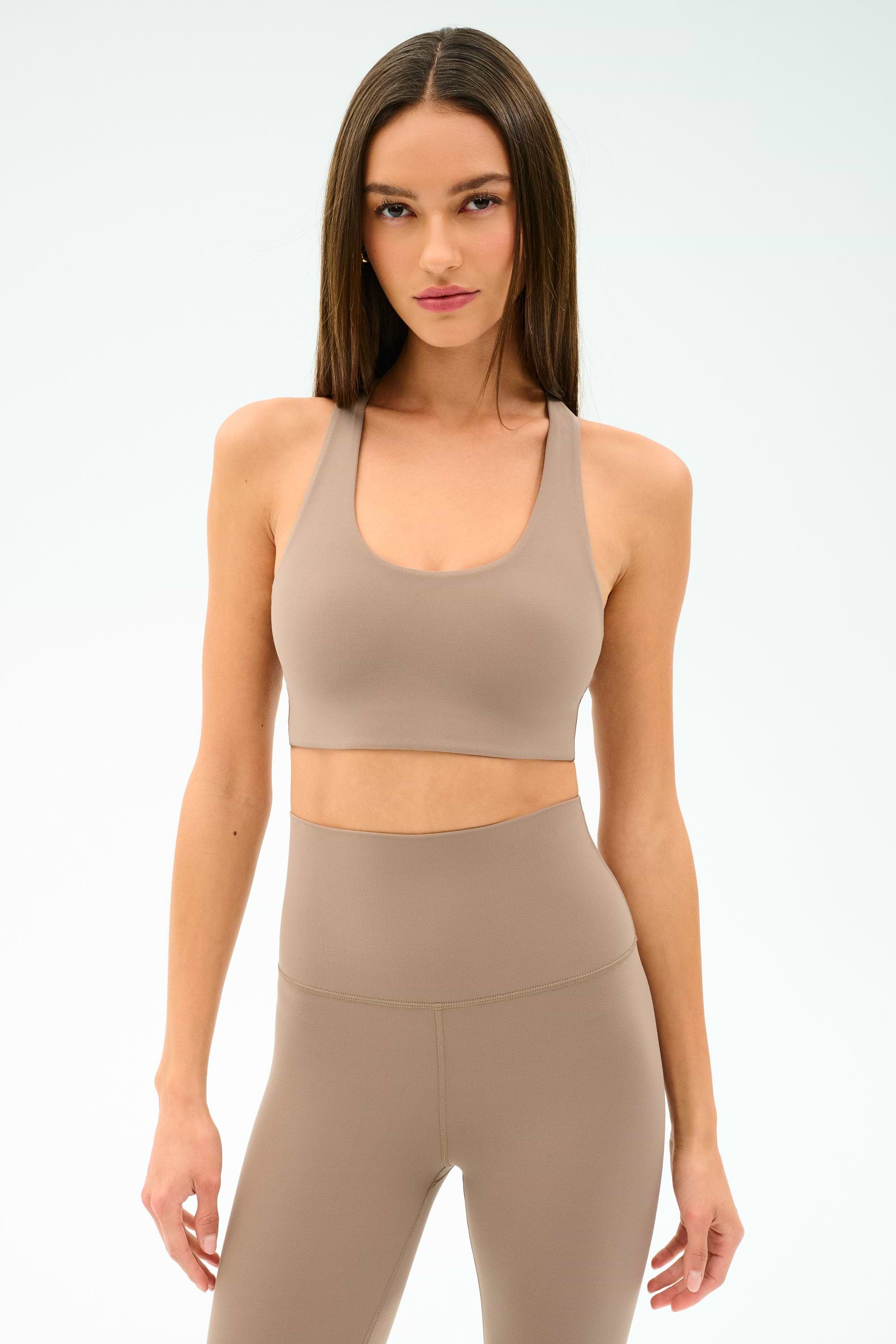 A woman models the SPLITS59 Sara Airweight Bra in Latte with matching high-waisted leggings against a plain white background—ideal for hot yoga or Pilates.