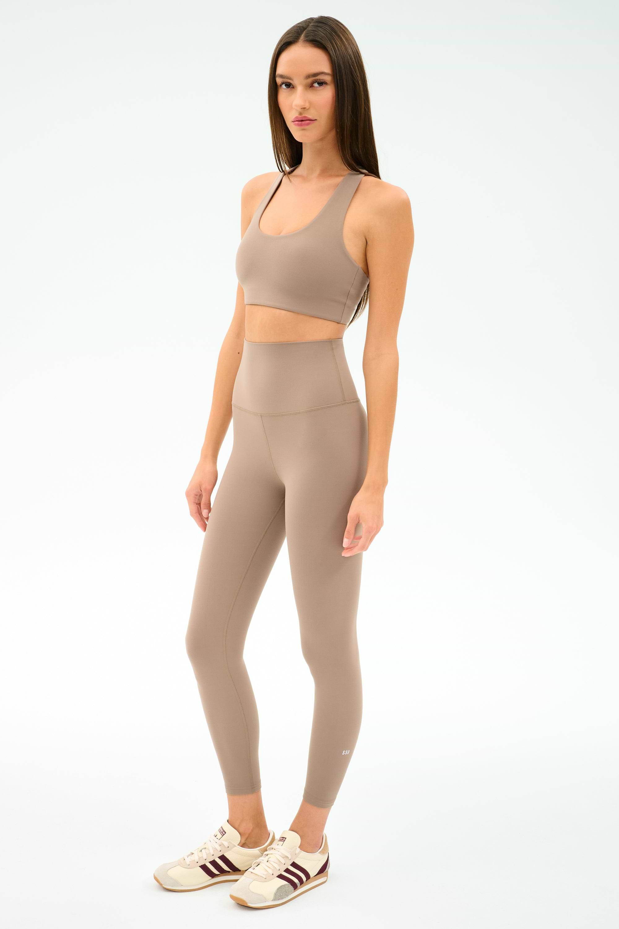 A woman poses against a white background wearing the SPLITS59 Airweight Super High Waist 7/8 leggings in Latte, paired with a taupe sports bra and beige sneakers featuring maroon stripes—an ideal outfit for hot yoga.