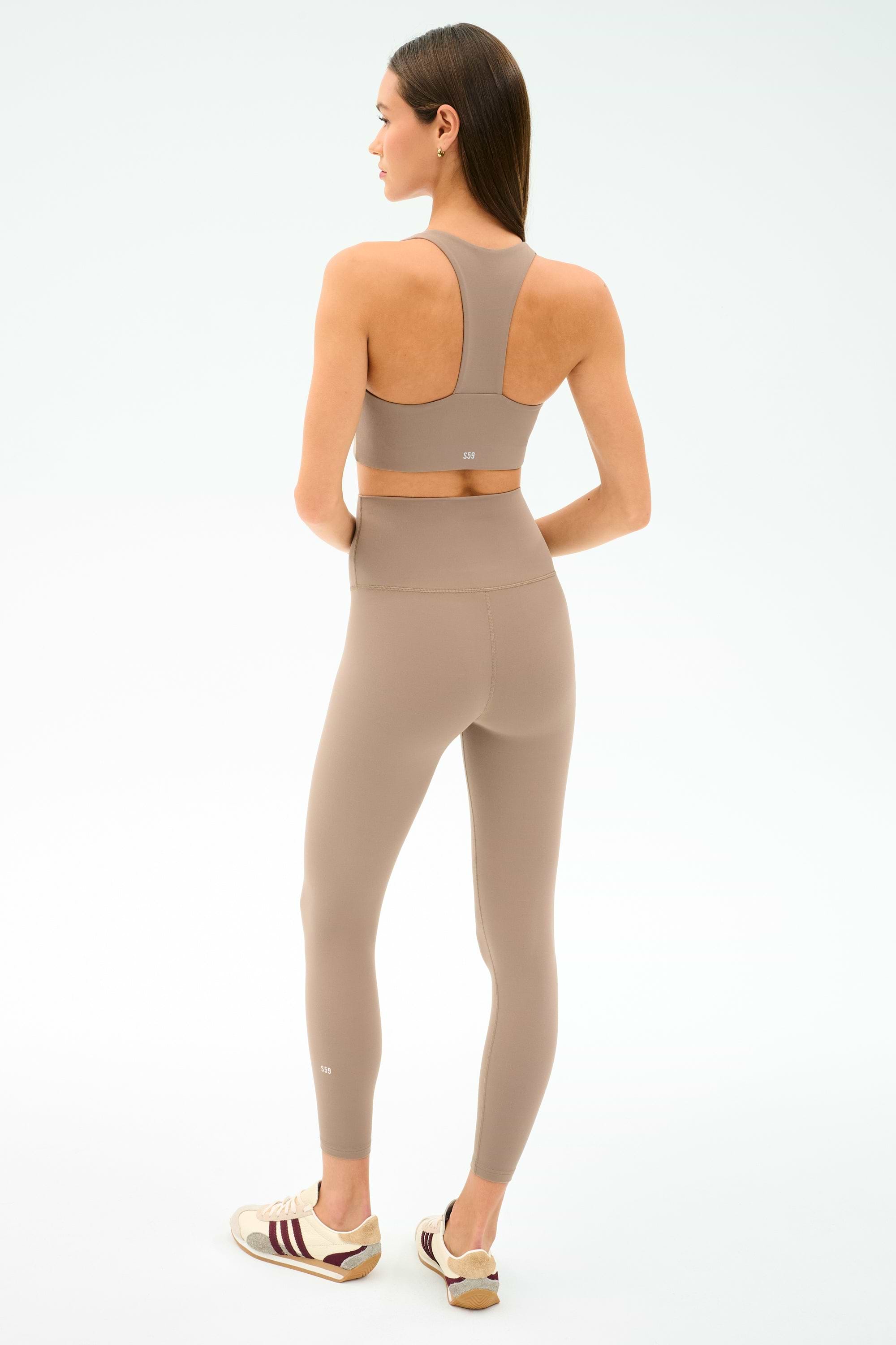 A woman stands facing away in the SPLITS59 Sara Airweight Bra - Latte and matching high-waisted leggings—ideal for Pilates or hot yoga—with beige sneakers, against a plain light background.