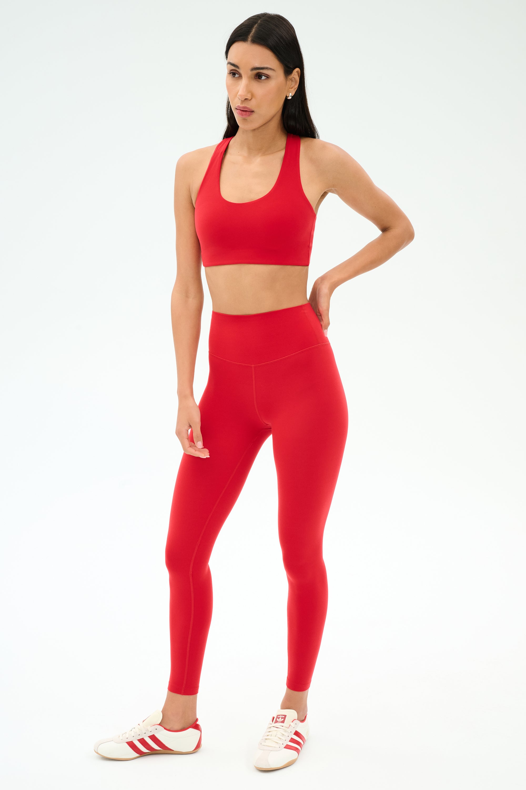 A woman stands against a white background wearing the SPLITS59 Sara Airweight Bra in Pirate Red with matching leggings, ideal for hot yoga or Pilates, paired with white sneakers accented by red stripes.
