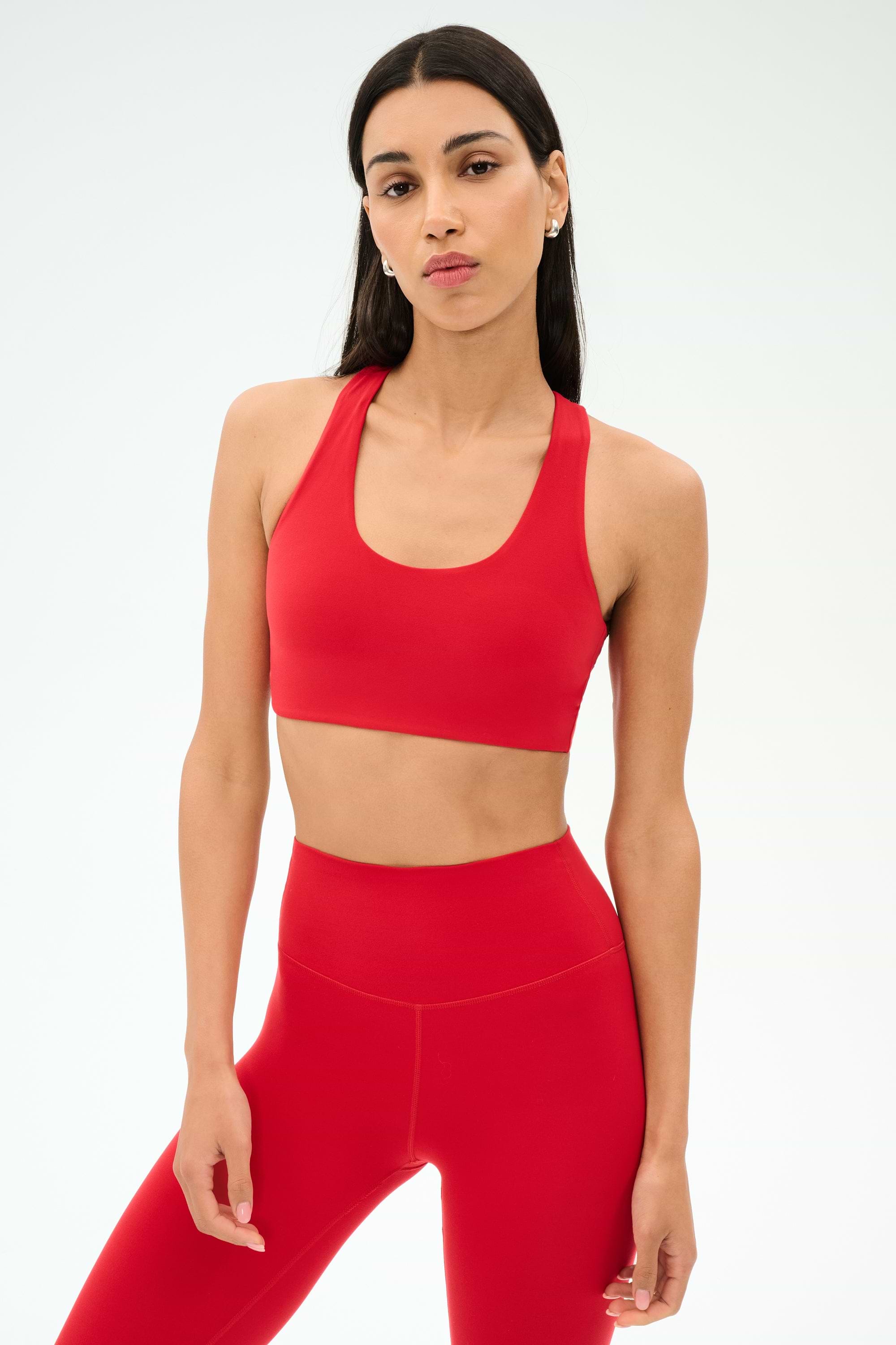A woman with long dark hair wears the SPLITS59 Sara Airweight Bra in Pirate Red and matching high-waisted leggings against a plain light background—ideal for hot yoga or Pilates.