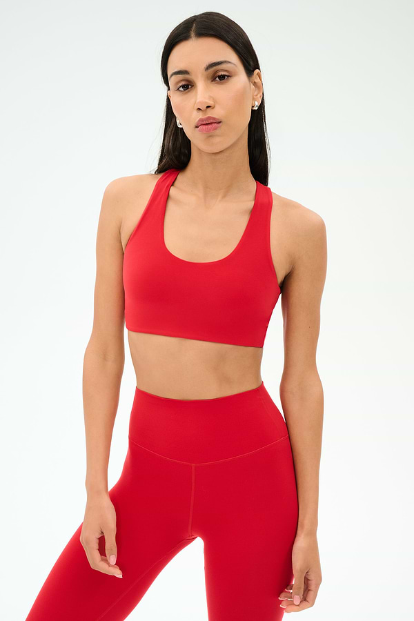 A woman with long dark hair wears the SPLITS59 Sara Airweight Bra in Pirate Red and matching high-waisted leggings against a plain light background—ideal for hot yoga or Pilates.