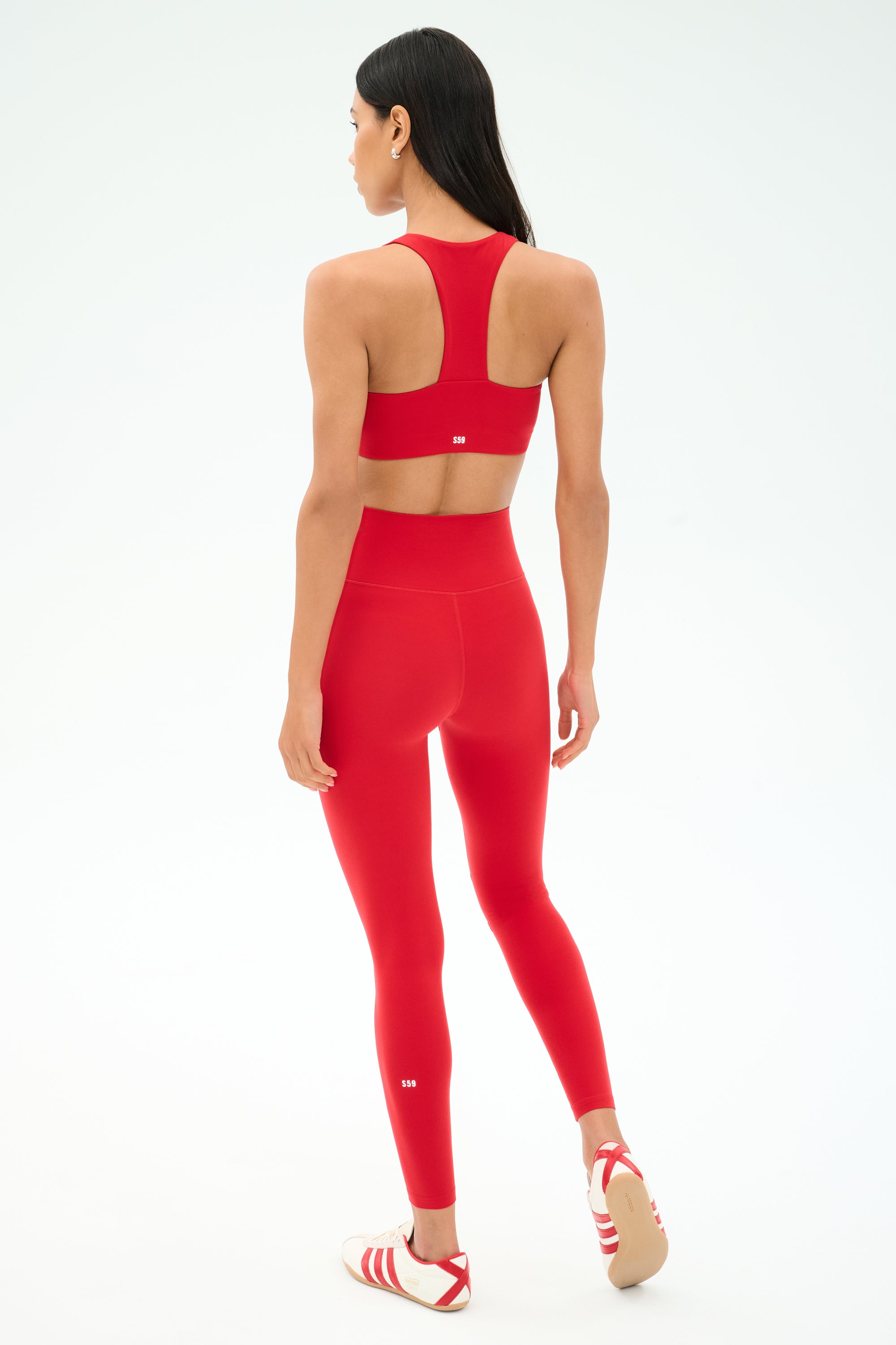 A woman stands facing away, wearing a matching SPLITS59 Sara Airweight Bra in Pirate Red and leggings with white sandals—perfect for hot yoga or Pilates. The background is plain white.