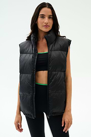 A woman wears the SPLITS59 Summit Nylon Puffer Vest in black over a sports bra and leggings, standing against a plain light background—the perfect lightweight layer for cool weather workouts.