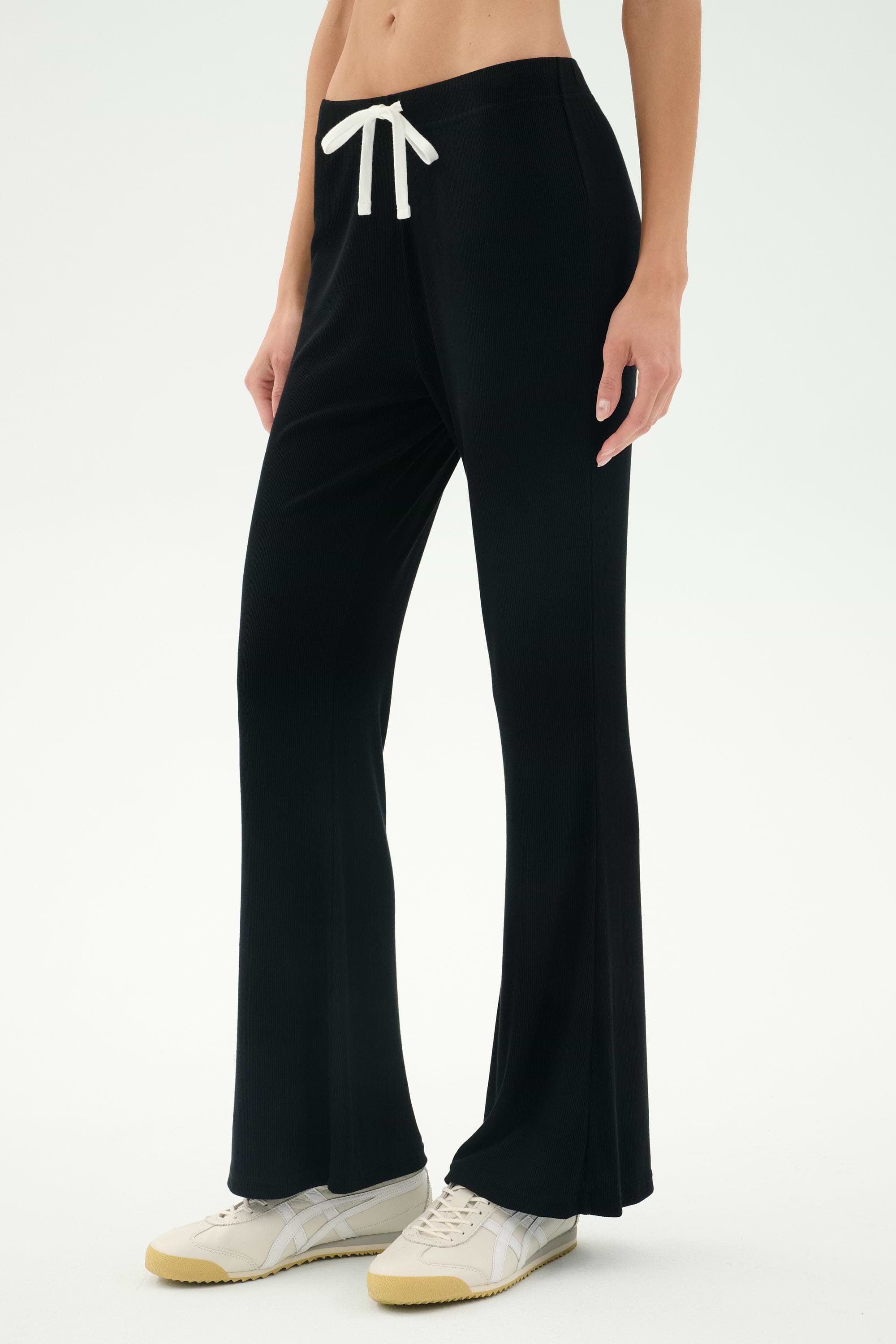 Someone wearing the SPLITS59 Blaire Rib Pant in black, which features a white drawstring, paired with white sneakers and standing against a plain light background.