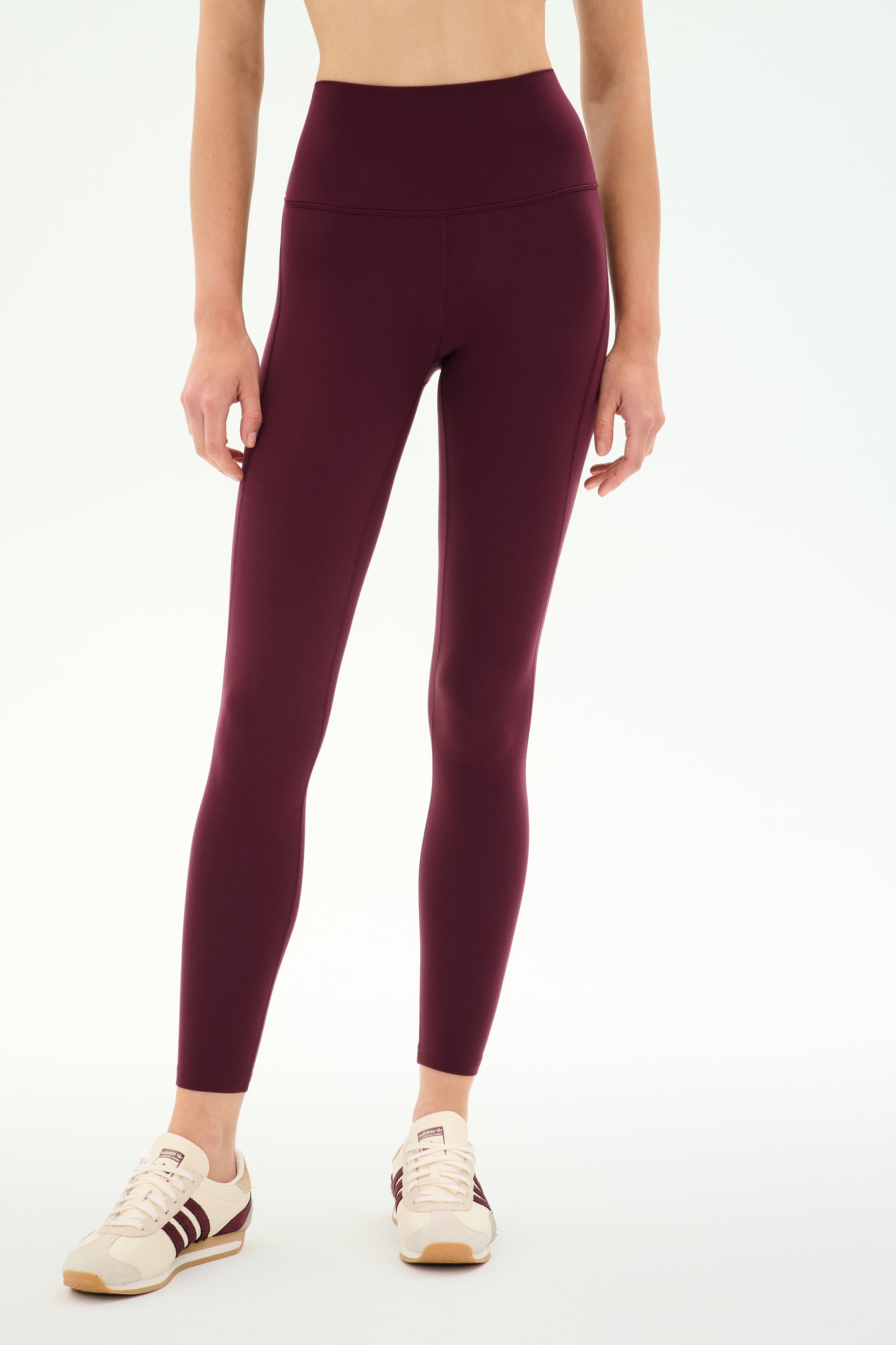 A woman stands against a plain white background, wearing SPLITS59’s Airweight High Waist 7/8 Pocket Legging in Dark Cherry and beige sneakers with maroon stripes.