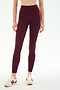 A woman stands against a plain white background, wearing SPLITS59’s Airweight High Waist 7/8 Pocket Legging in Dark Cherry and beige sneakers with maroon stripes.