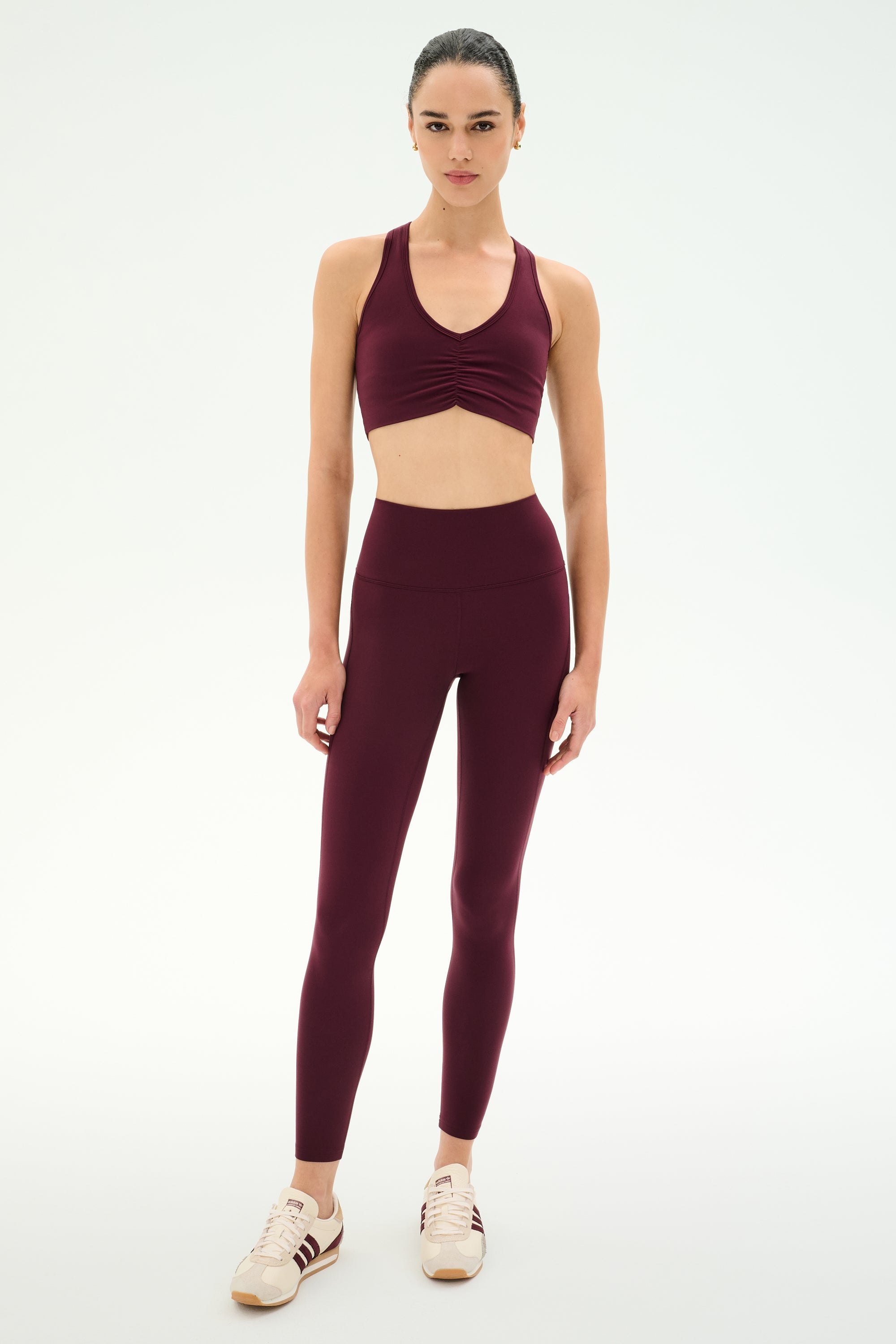A woman stands against a plain white background with a neutral expression, wearing the SPLITS59 Airweight Bra With Ruching in Dark Cherry, paired with matching high-waisted leggings crafted from stretch fabric.