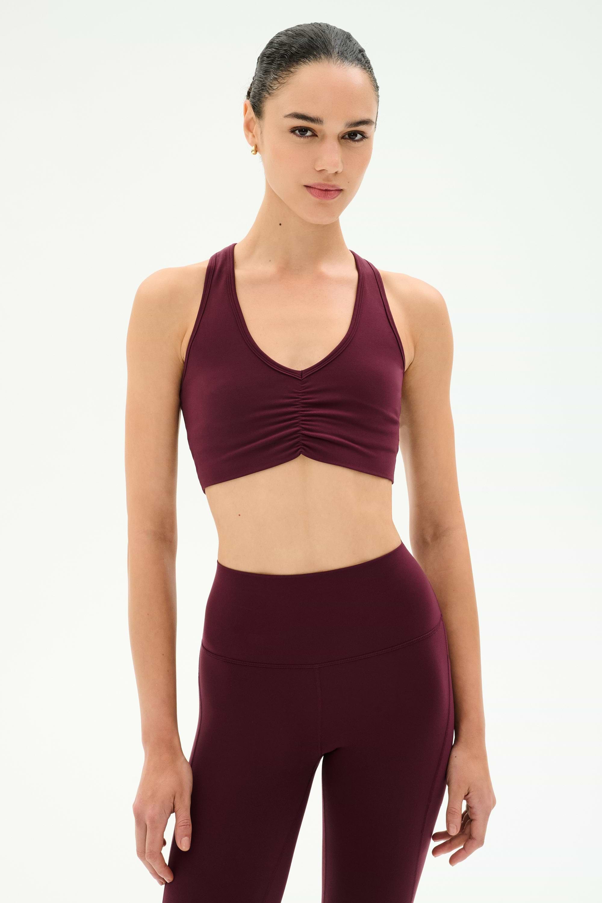 A woman wears the SPLITS59 Airweight Bra With Ruching in Dark Cherry, paired with matching high-waisted leggings, both crafted from soft stretch fabric for ultimate comfort, standing against a plain white background.
