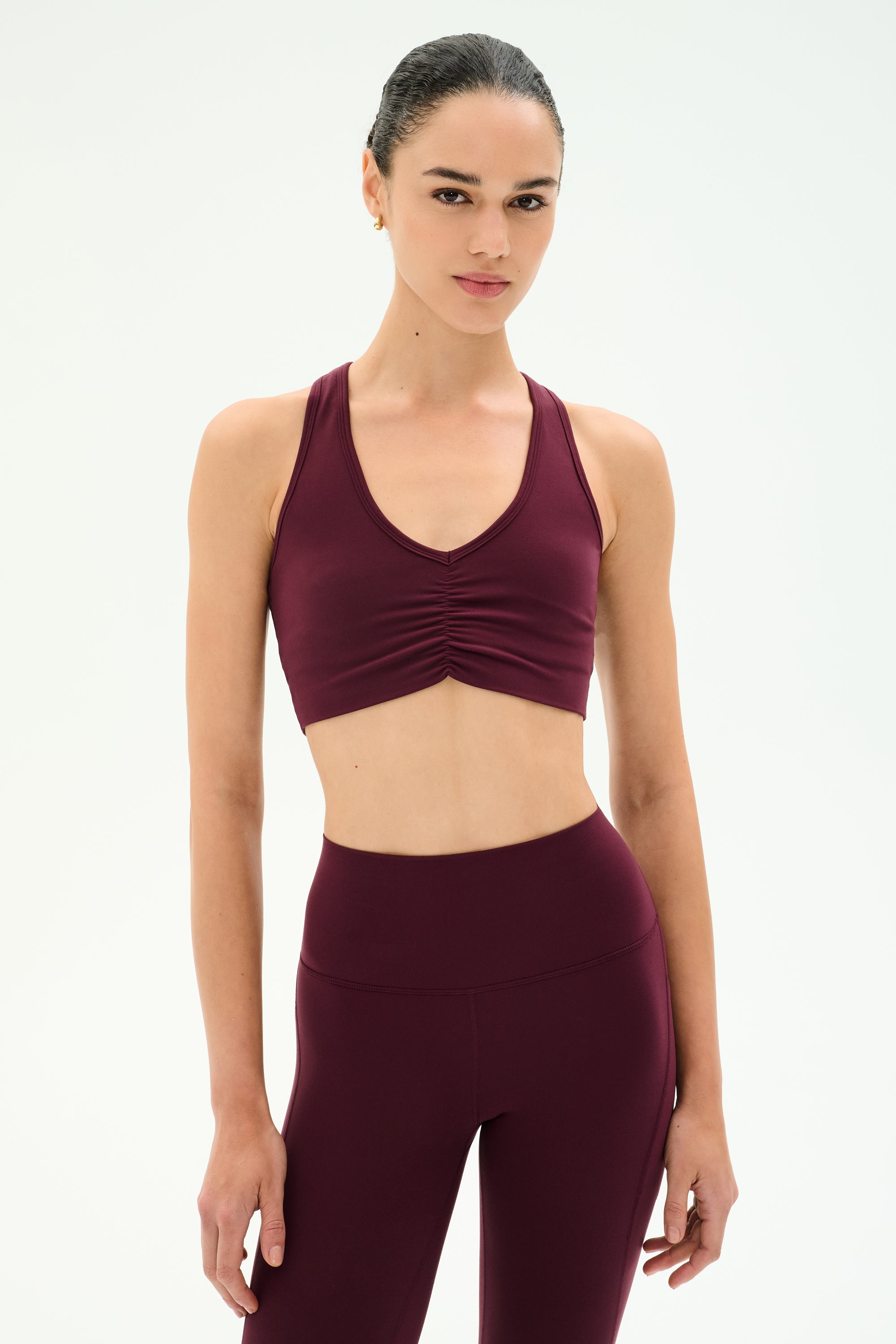 A woman wears the SPLITS59 Airweight Bra With Ruching in Dark Cherry, paired with matching high-waisted leggings, both crafted from soft stretch fabric for ultimate comfort, standing against a plain white background.