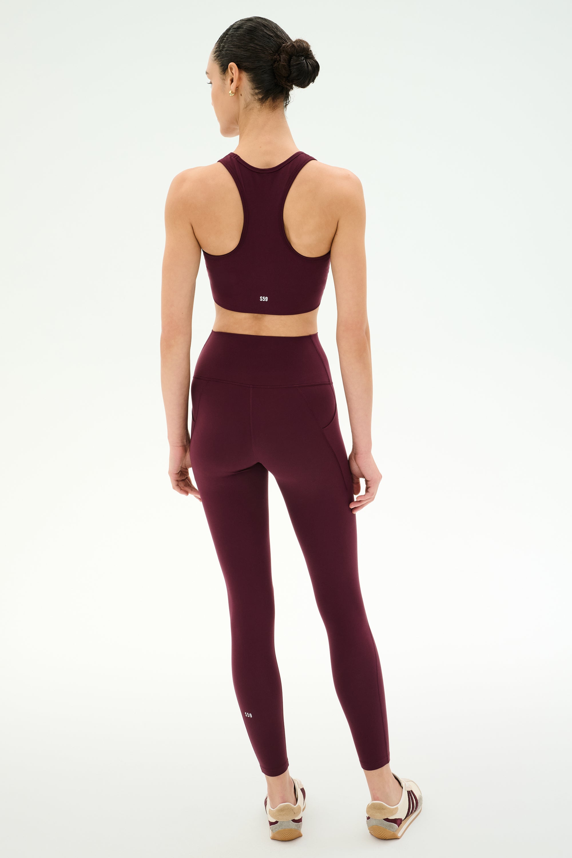 A woman stands facing away on a plain white background, wearing the SPLITS59 Airweight High Waist 7/8 Pocket Legging in Dark Cherry and a matching burgundy sports bra, paired with beige sneakers.