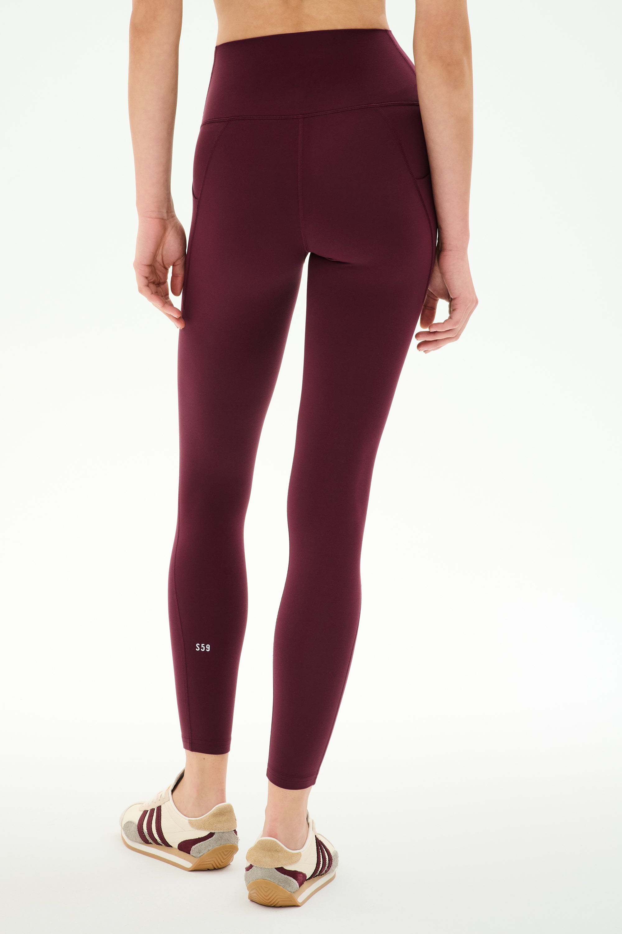 A person is shown from the back, standing against a plain white background, wearing the SPLITS59 Airweight High Waist 7/8 Pocket Legging in Dark Cherry and beige sneakers with maroon stripes.