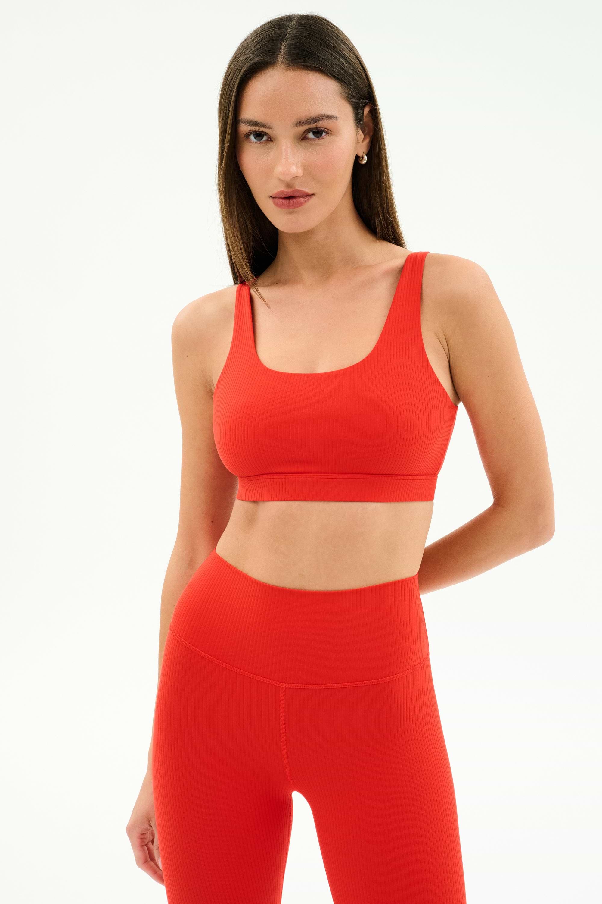 A woman stands against a white background wearing the SPLITS59 Airweight Rib Bra in Lotus Red, paired with high-waisted leggings—an ideal outfit for hot yoga.