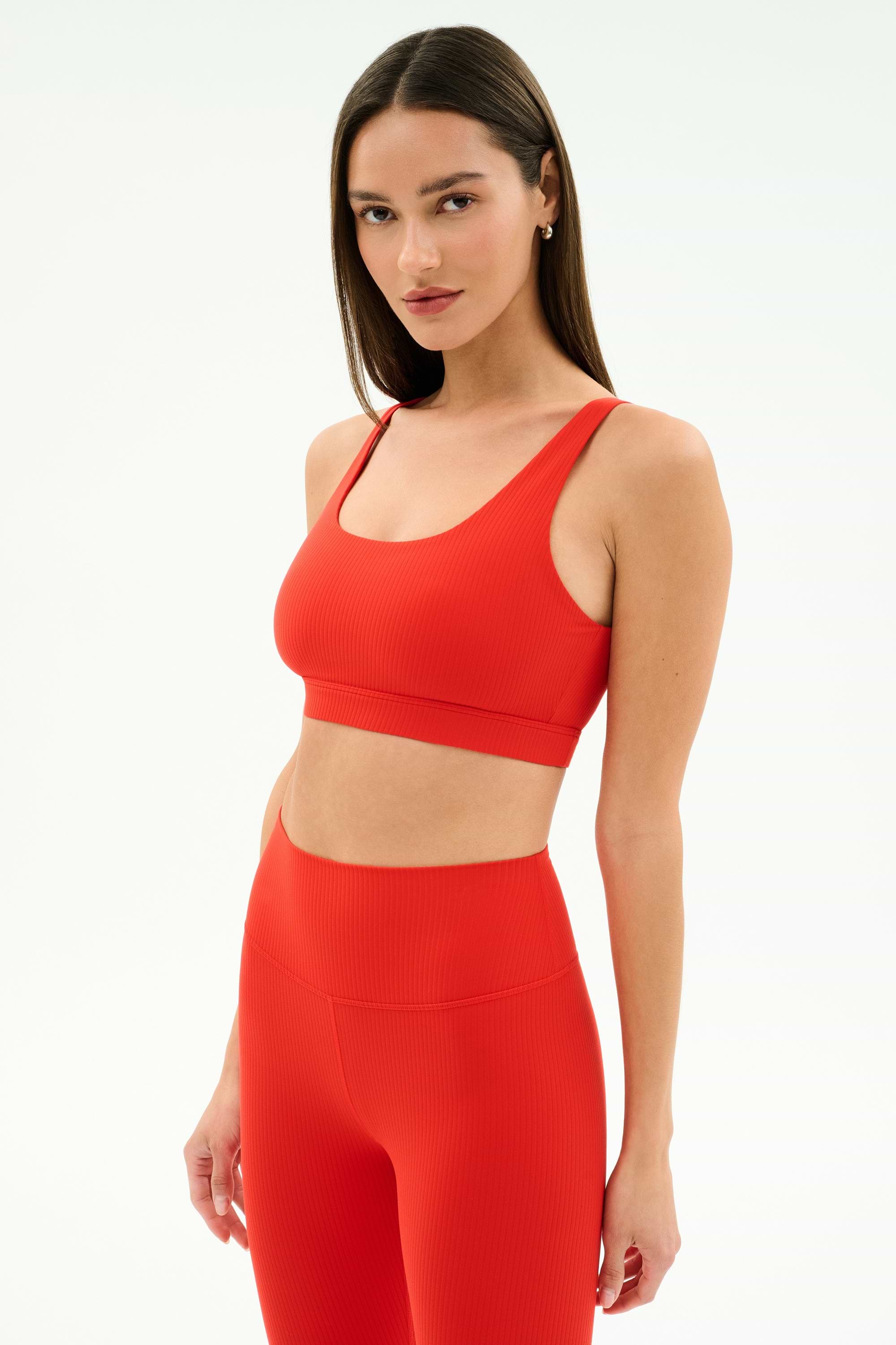 A woman wears the SPLITS59 Airweight Rib Bra in Lotus Red with matching high-waisted leggings, made from ribbed airweight fabric—ideal for hot yoga—posing against a plain white background.