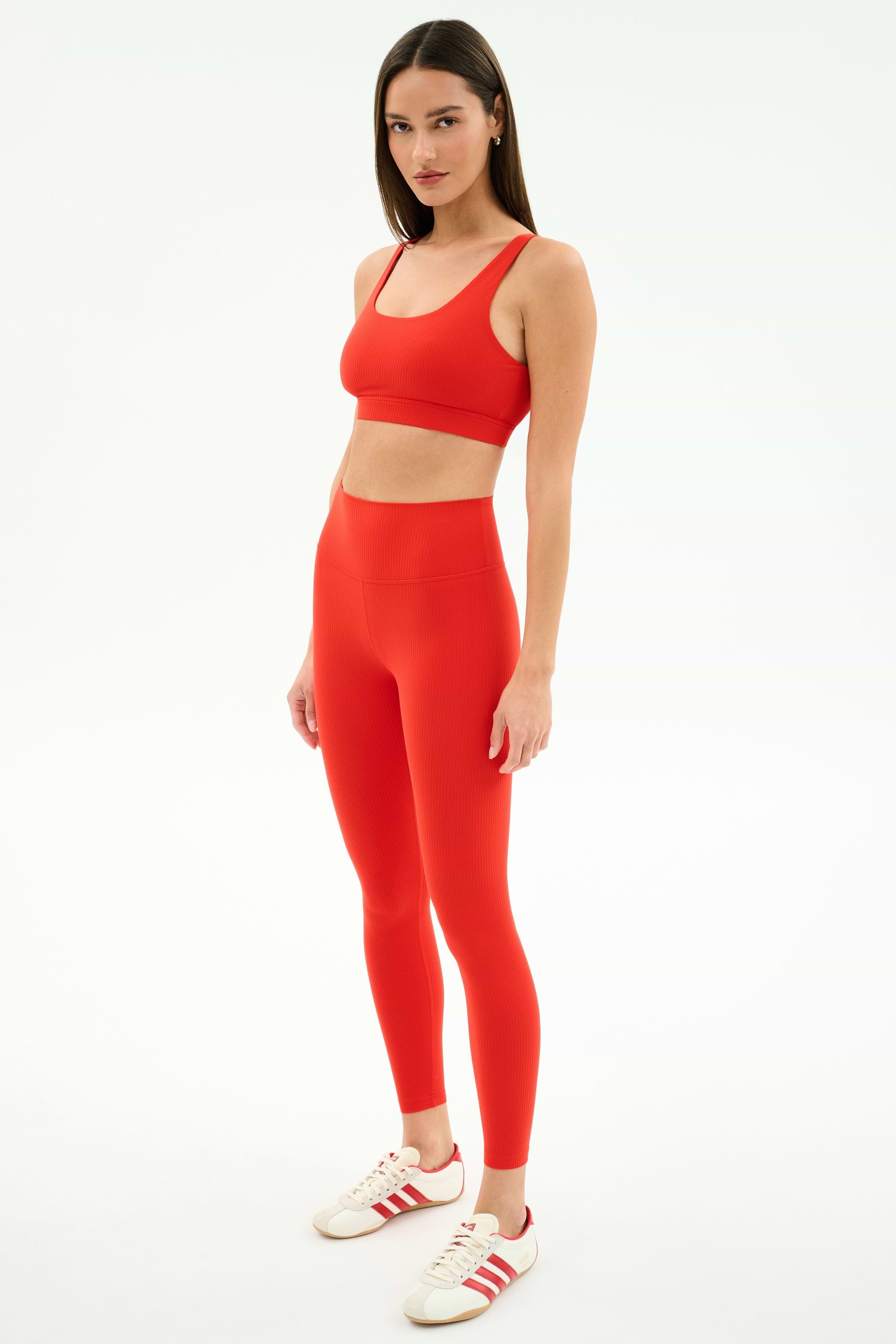 A woman poses against a plain background in the SPLITS59 Airweight Rib Bra - Lotus Red, paired with matching leggings and white sneakers with red stripes—an ideal set for hot yoga.