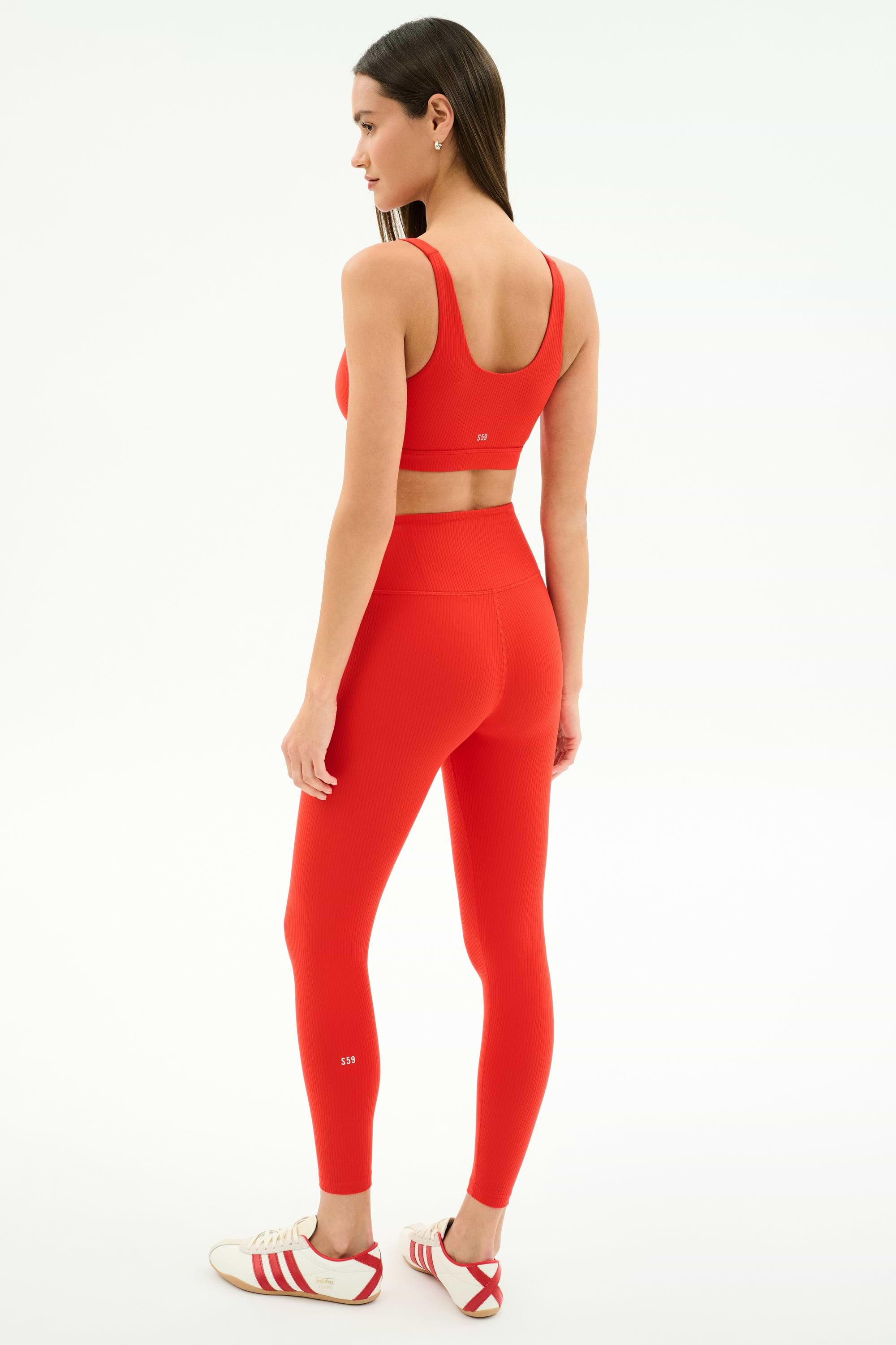 A woman, facing away and looking to the side, wears SPLITS59 Airweight Rib High Waist 7/8 leggings in Lotus Red with a matching sports bra and white Adidas sneakers against a plain white background.