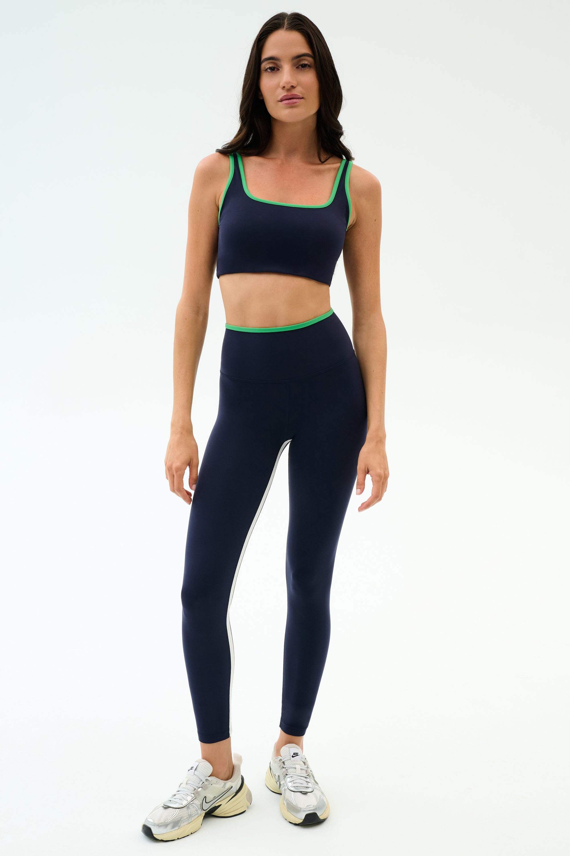 A woman stands against a plain background wearing the SPLITS59 Cait Rigor Bra in Indigo/Grass and matching leggings, made from Rigor fabric for high-impact workouts, styled with white sneakers.