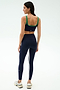 A woman faces away, wearing the SPLITS59 Cait Rigor Bra in Indigo/Grass and matching navy leggings with a white side stripe, paired with white sneakers against a plain light background.