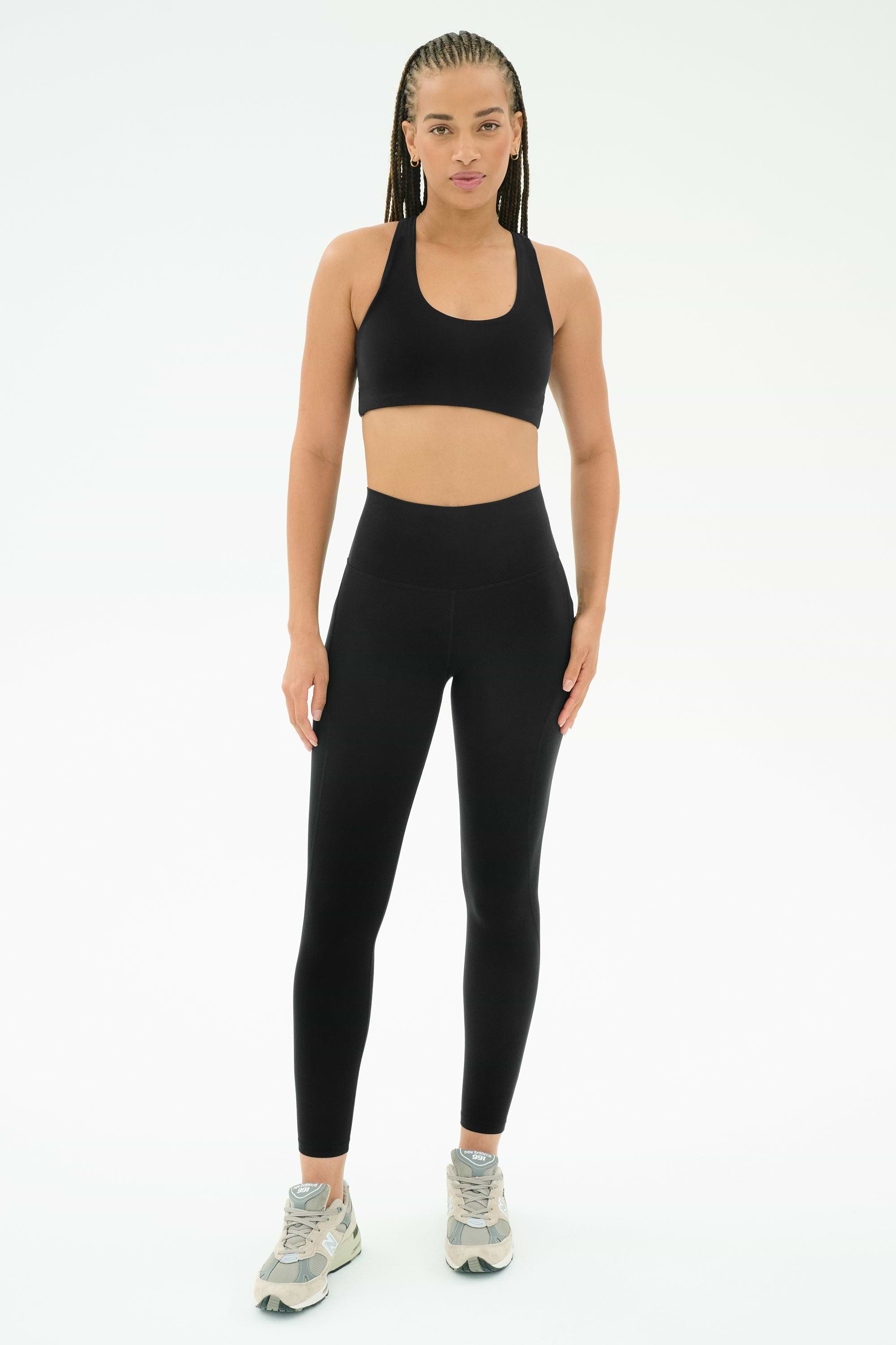 A person faces forward in SPLITS59's Airweight High Waist 7/8 Pocket Legging - Black, a black sports bra, and gray athletic shoes, pictured against a plain white background.